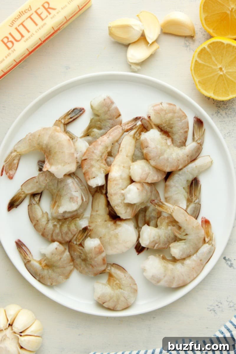 Butter, garlic cloves, lemon and raw shrimp on a plate, set on a gray board.