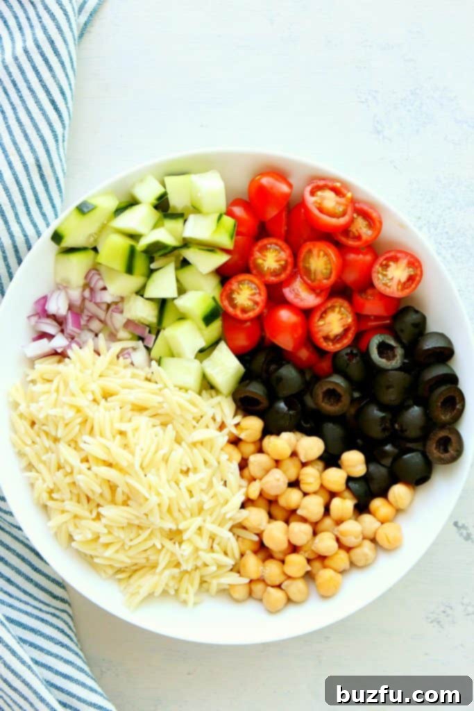 Fresh ingredients for Orzo Pasta Salad including cooked orzo, cherry tomatoes, cucumber, olives, and chickpeas in bowls.