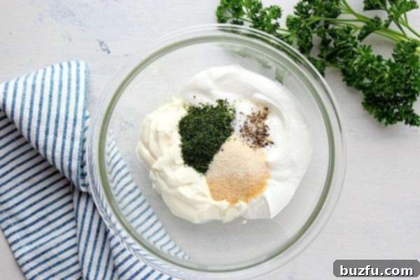 A glass bowl filled with the creamy mixture of mayonnaise, sour cream, and various seasonings, freshly mixed and ready for the next step.
