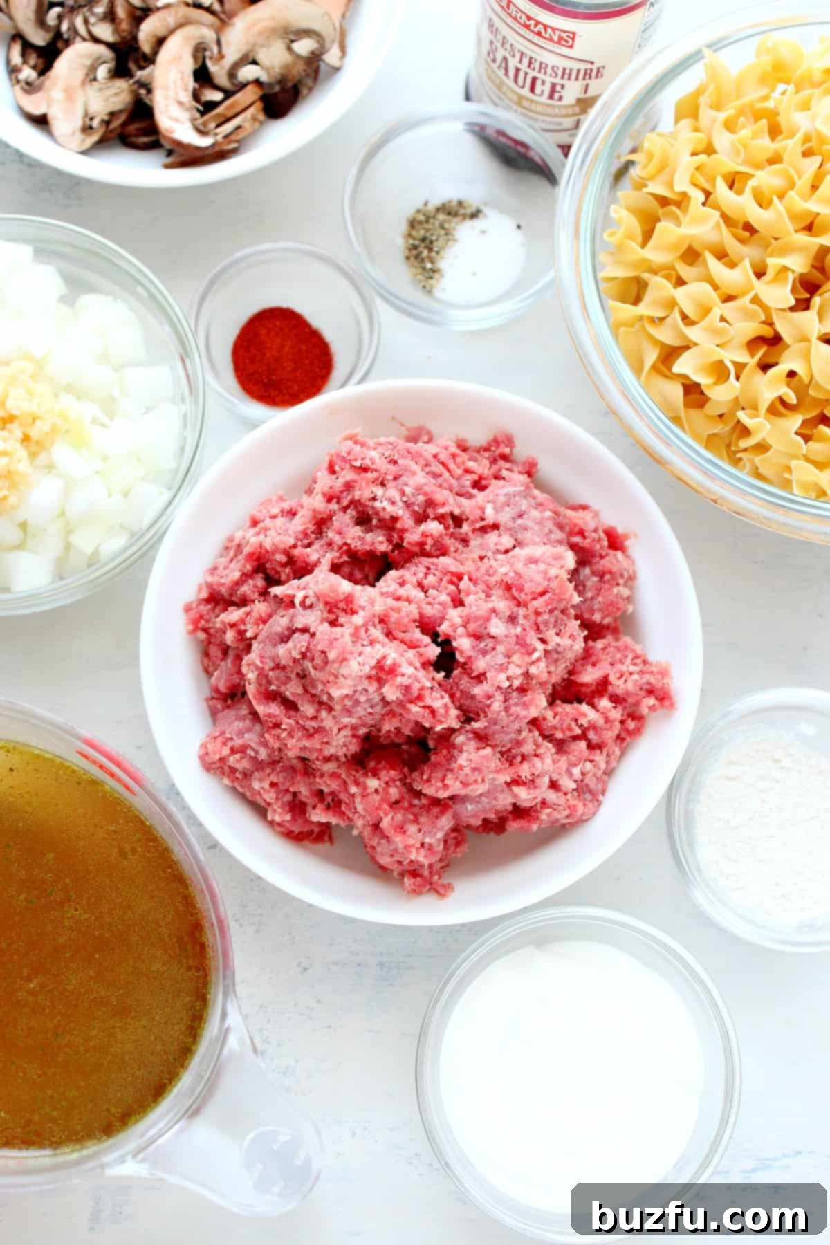 Instant Pot Beef Stroganoff ingredients including ground beef, mushrooms, wide egg noodles, beef broth, and sour cream laid out on a white cutting board.