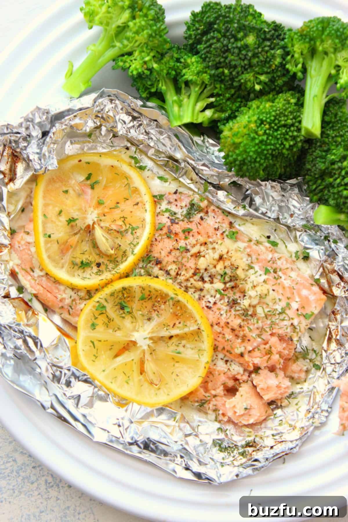 Freshly baked salmon in foil, garnished with lemon slices and herbs, on a white plate ready to be served.