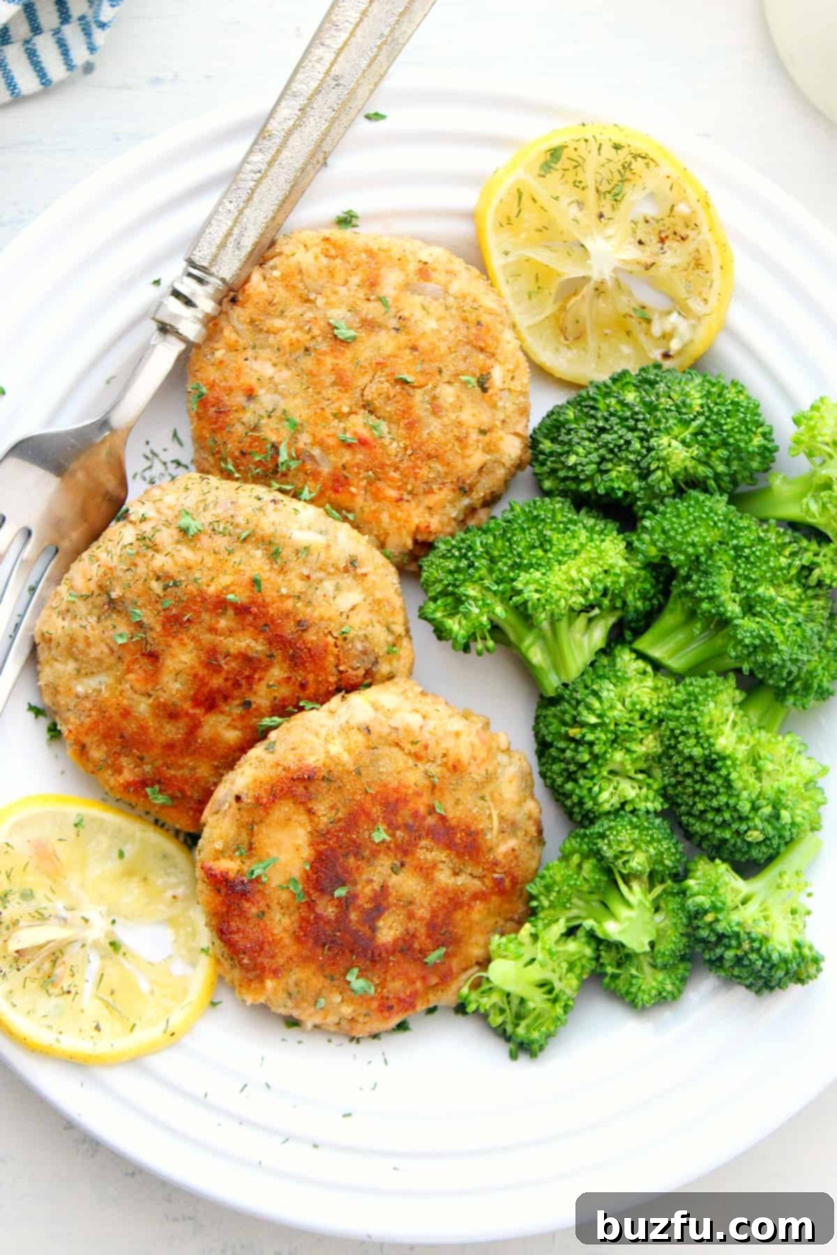 Easy Salmon Cakes served with healthy sides Two golden brown salmon cakes on a white plate with steamed broccoli and lemon wedges.