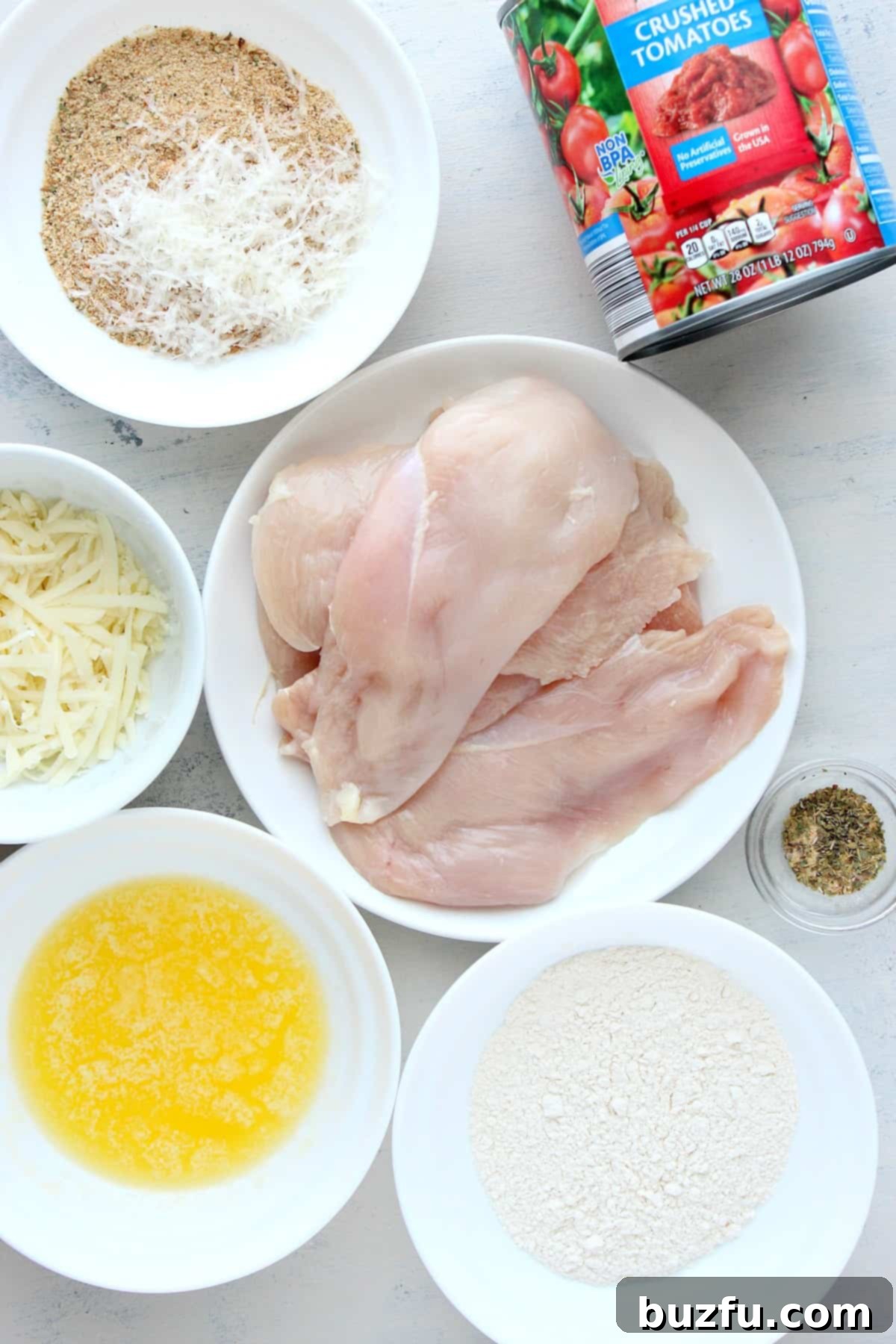 Various ingredients for chicken parmesan laid out in small bowls, including chicken breasts, breadcrumbs, cheese, and seasonings.