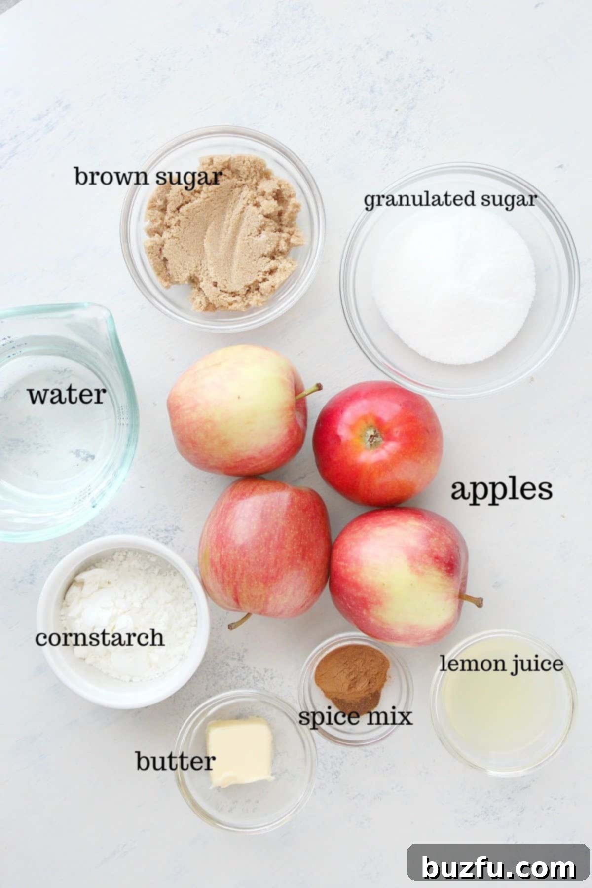 All the fresh ingredients for homemade apple pie filling laid out on a clean white cutting board, including apples, sugars, spices, and lemon juice.