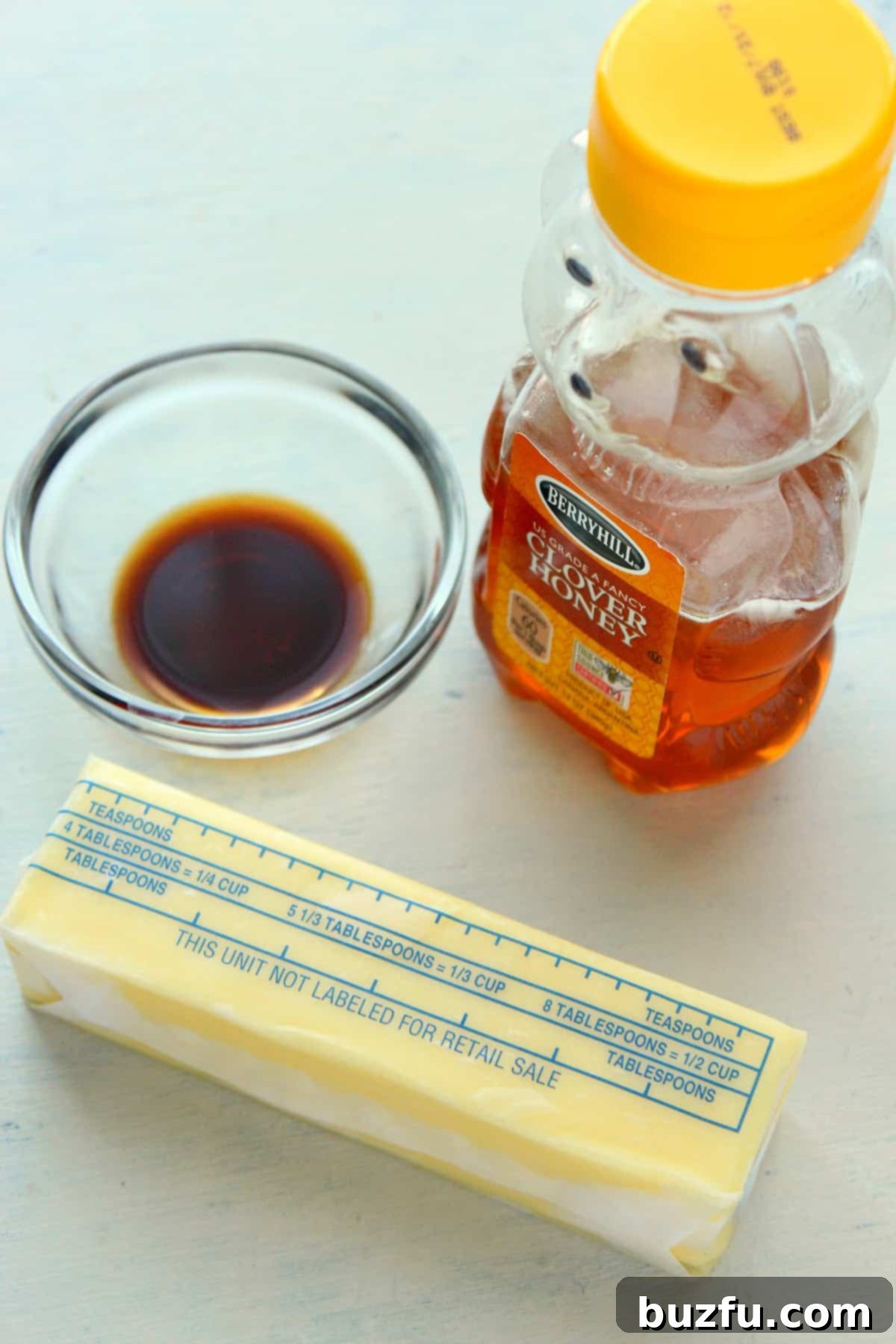Raw ingredients for homemade honey butter: a stick of unsalted butter, a jar of honey, and a bottle of vanilla extract are neatly arranged on a wooden cutting board.