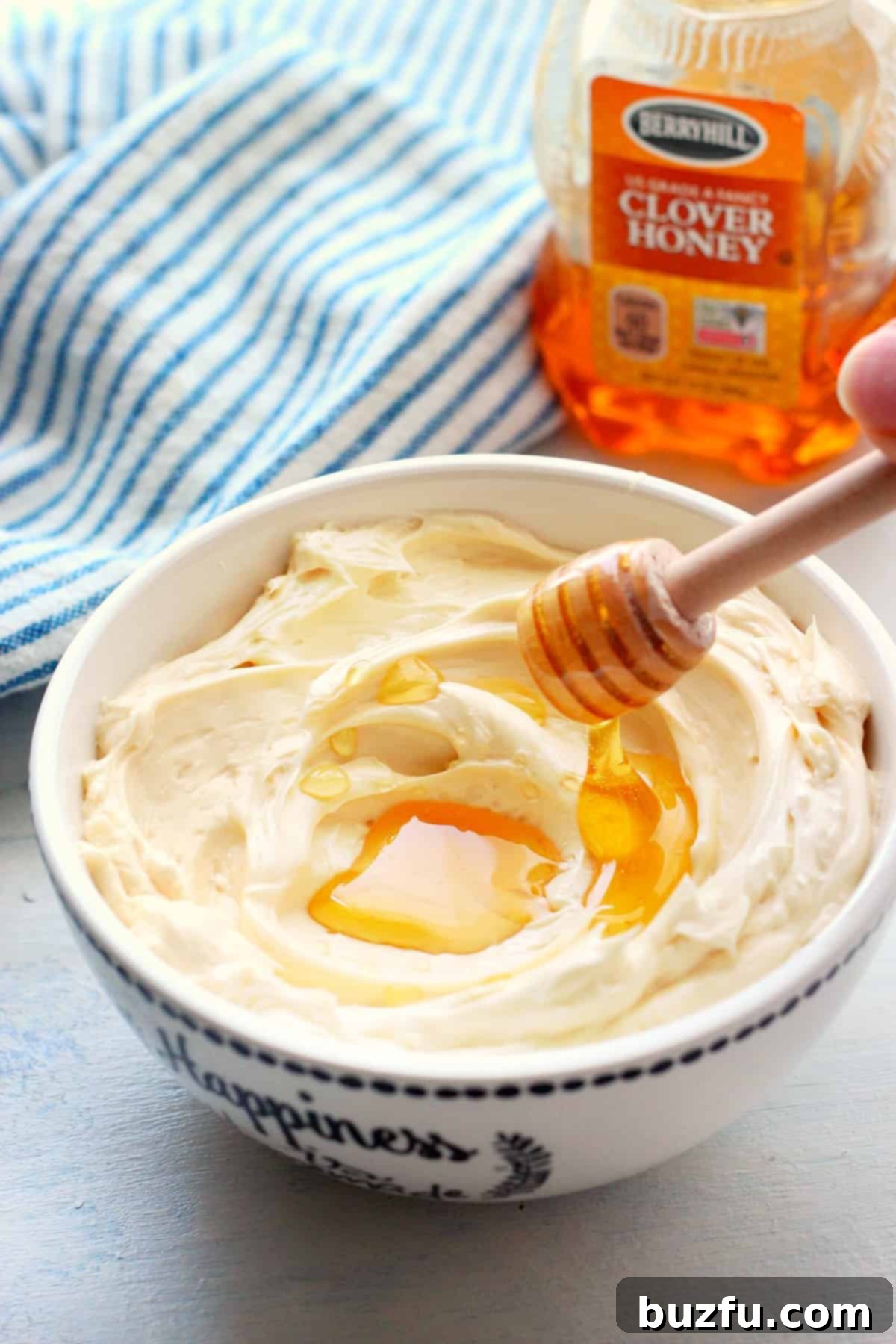 A large dollop of golden, whipped honey butter with a generous drizzle of honey cascading over it, presented on a clean white wooden board with a honey dipper nearby.