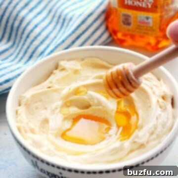 A dollop of fluffy honey butter with a drizzle of honey on a white board, next to a honey dipper.