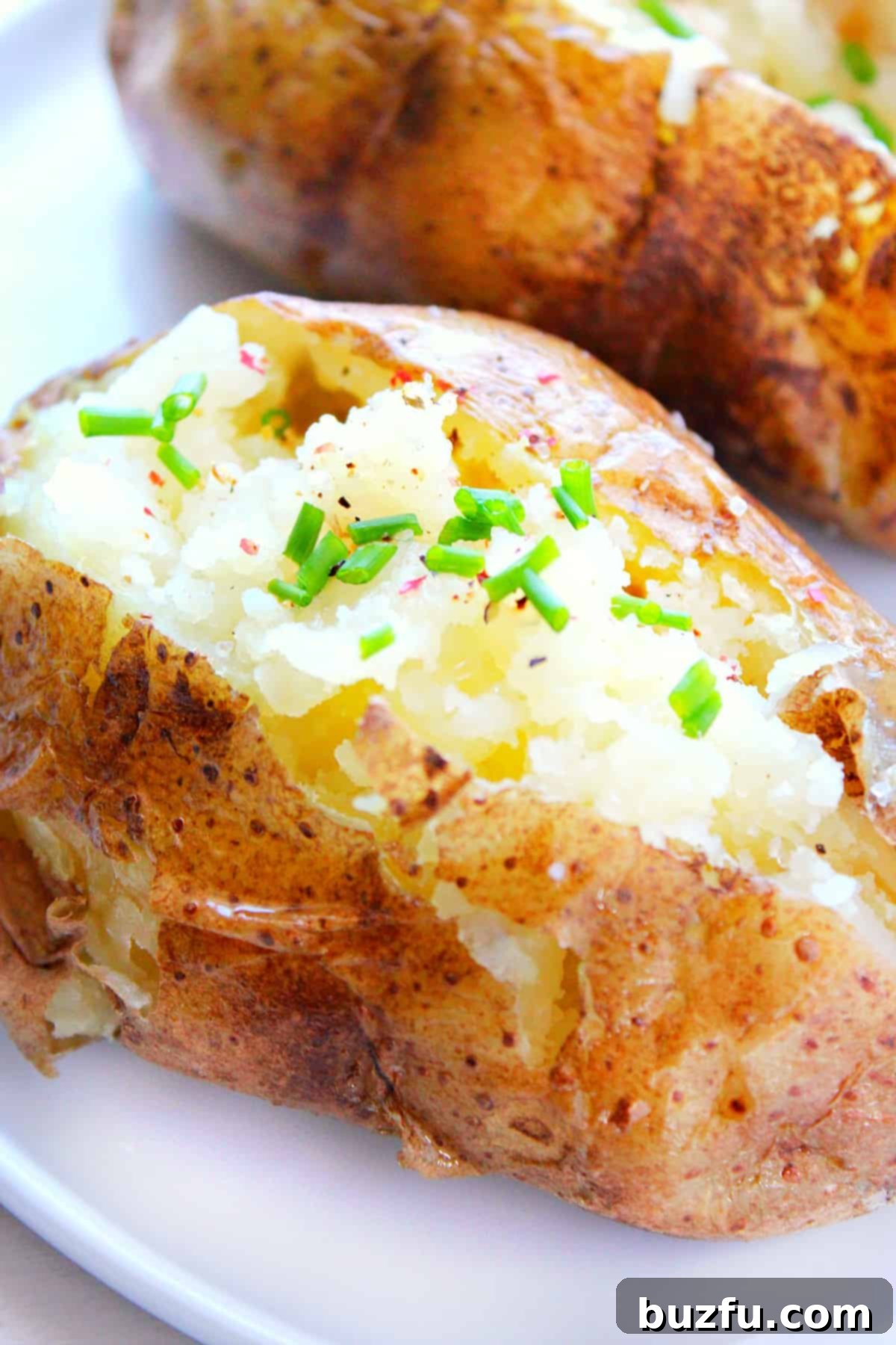 A perfectly baked potato topped generously with melting butter and freshly chopped chives, resting on a white plate.