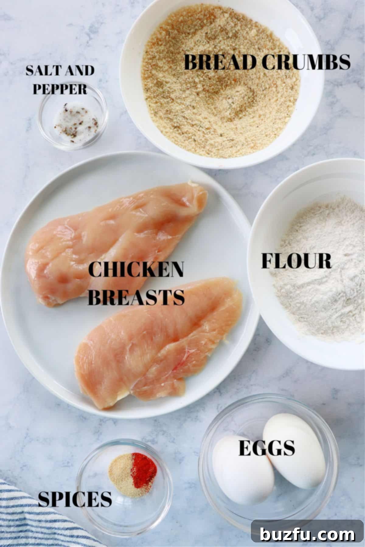 Breaded chicken cutlets ingredients preparation A selection of raw ingredients for breaded chicken cutlets laid out on a wooden cutting board, including chicken breasts, eggs, flour, bread crumbs, salt, pepper, garlic powder, and paprika.