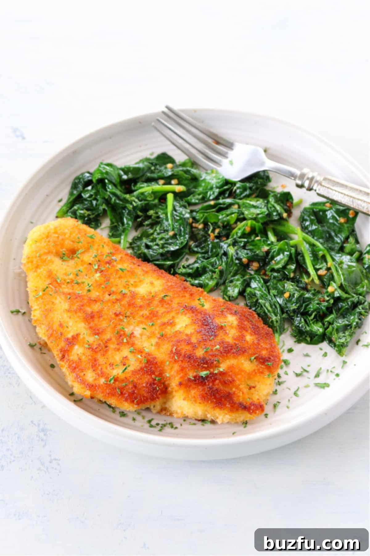 Serving crispy breaded chicken cutlets with sautéed spinach A golden brown breaded and fried chicken cutlet resting on a white plate alongside a generous portion of bright green sautéed spinach, garnished with a sprig of fresh parsley.