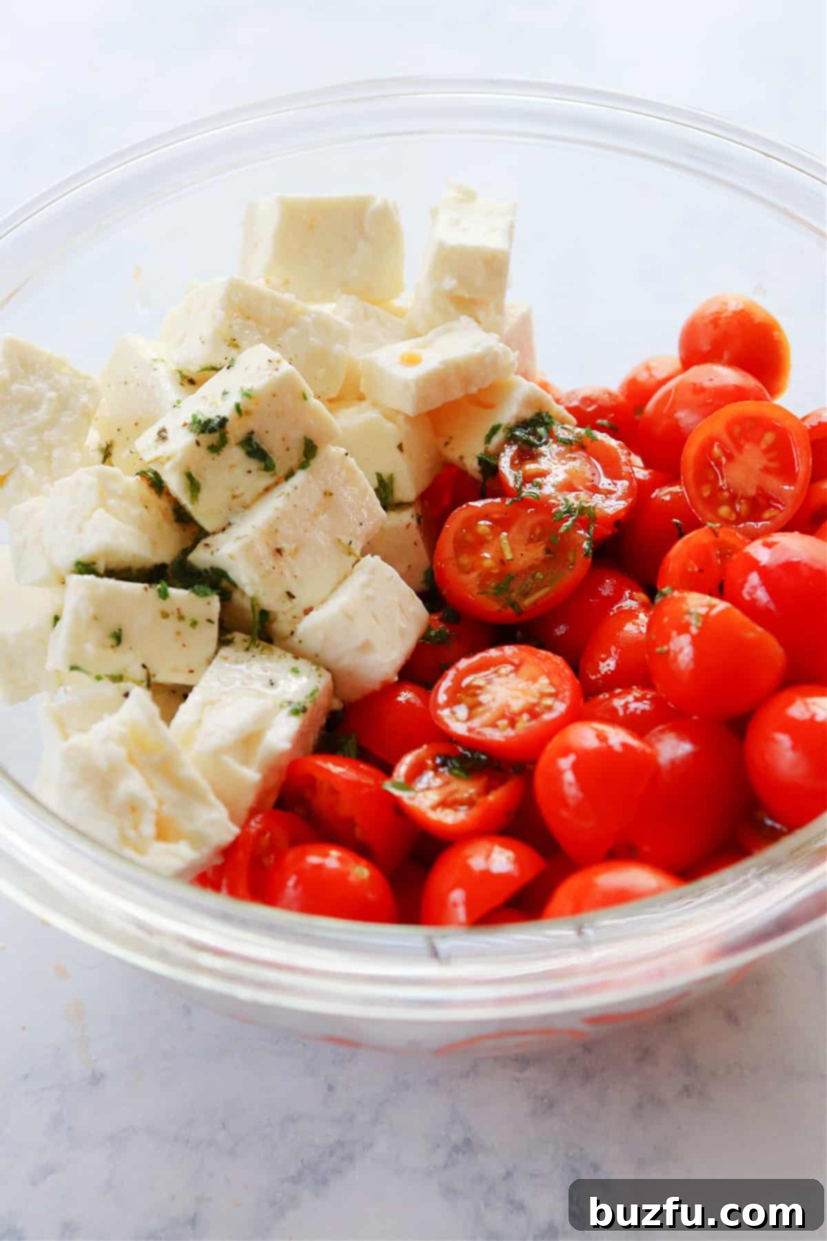 How to make tomato and feta salad step-by-step. Freshly cut feta cheese and halved cherry tomatoes tossed in a light dressing in a clear glass bowl, ready to be served.