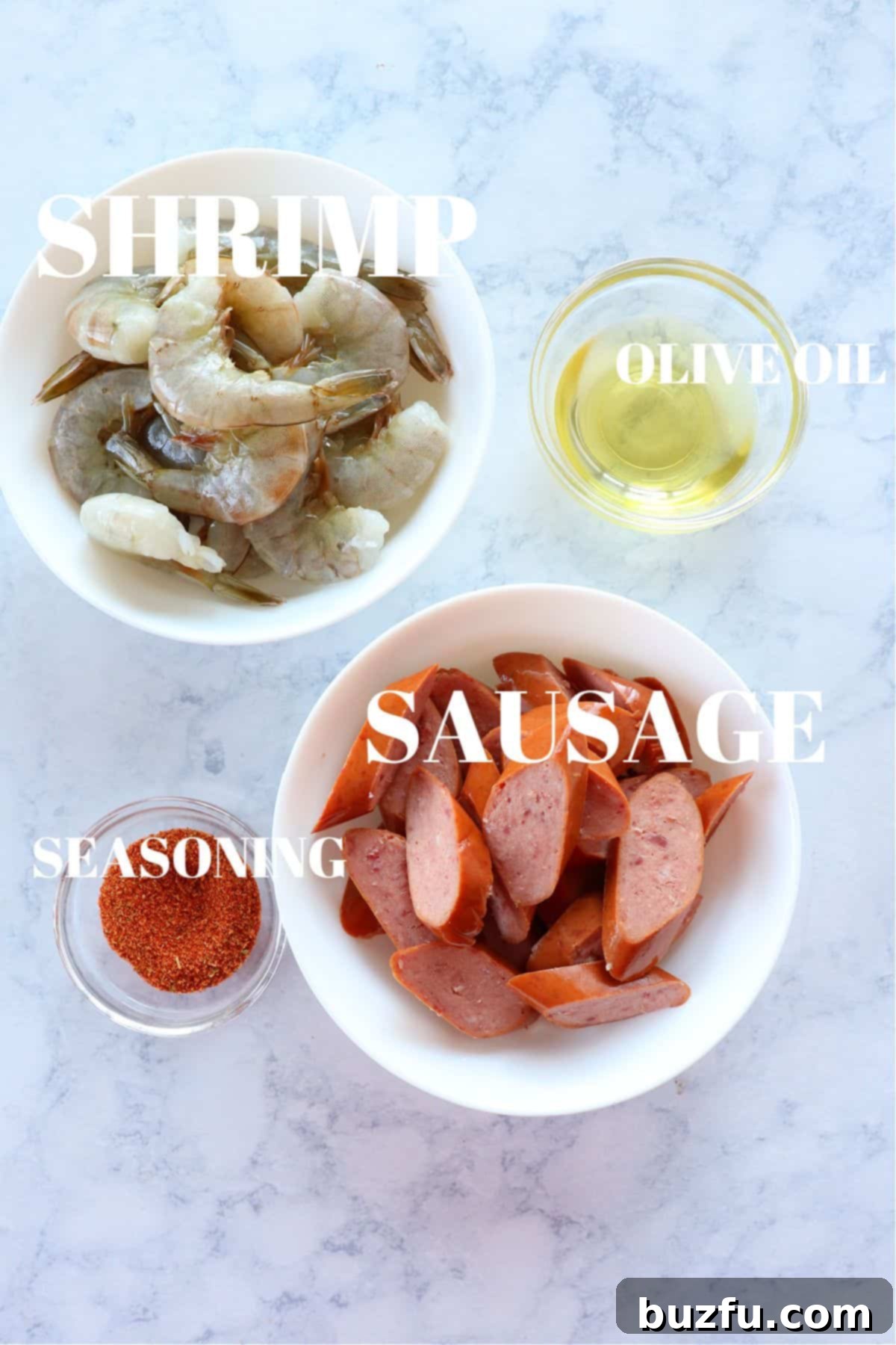 Fresh ingredients laid out on a marble board, including jumbo shrimp, smoked sausage, bell peppers, red onion, and olive oil.