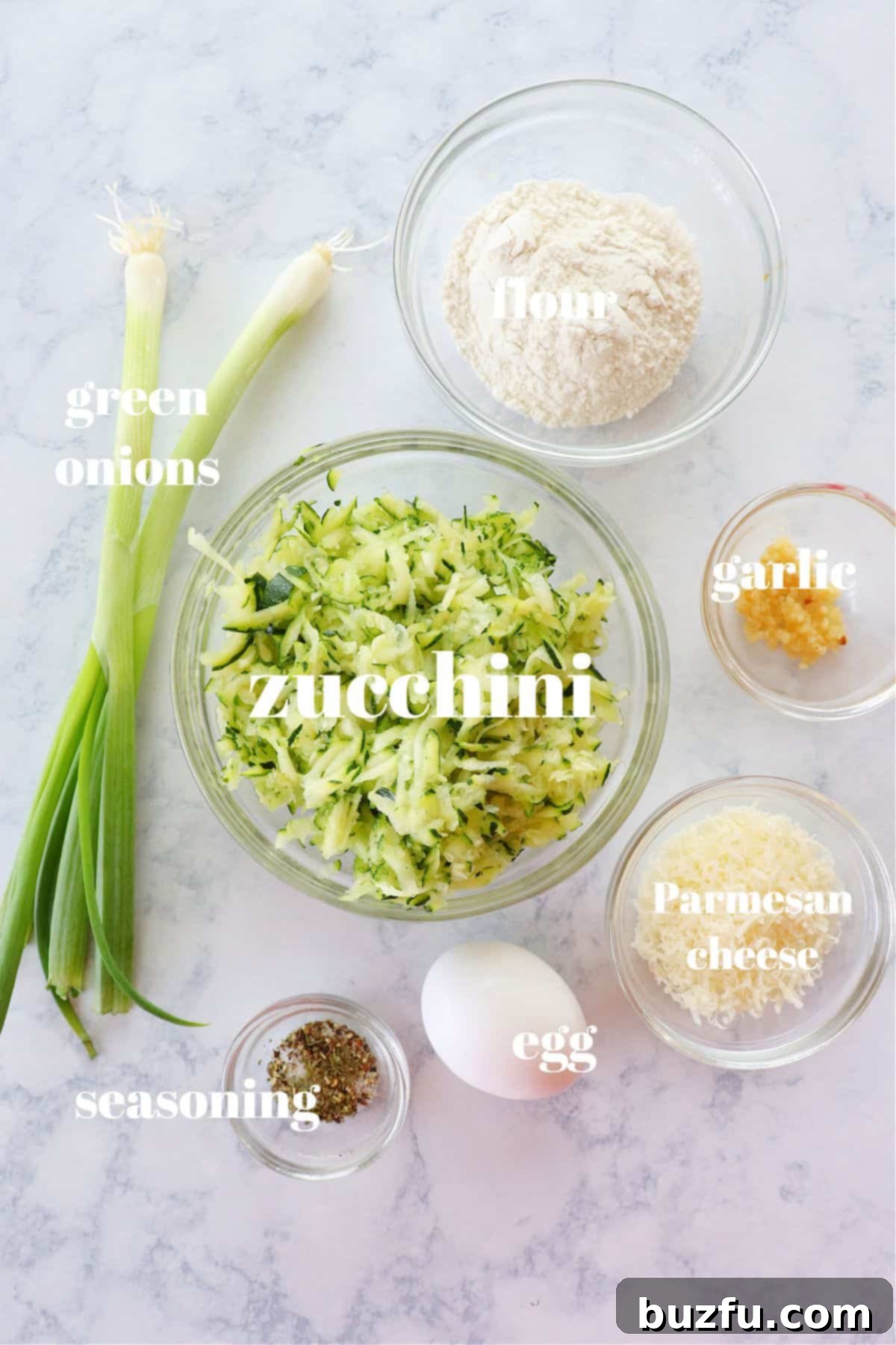 Zucchini Fritters Ingredients Layout A beautiful flat lay of fresh ingredients for zucchini fritters, including shredded zucchini, green onions, garlic, Parmesan cheese, flour, and an egg, on a white marble board.