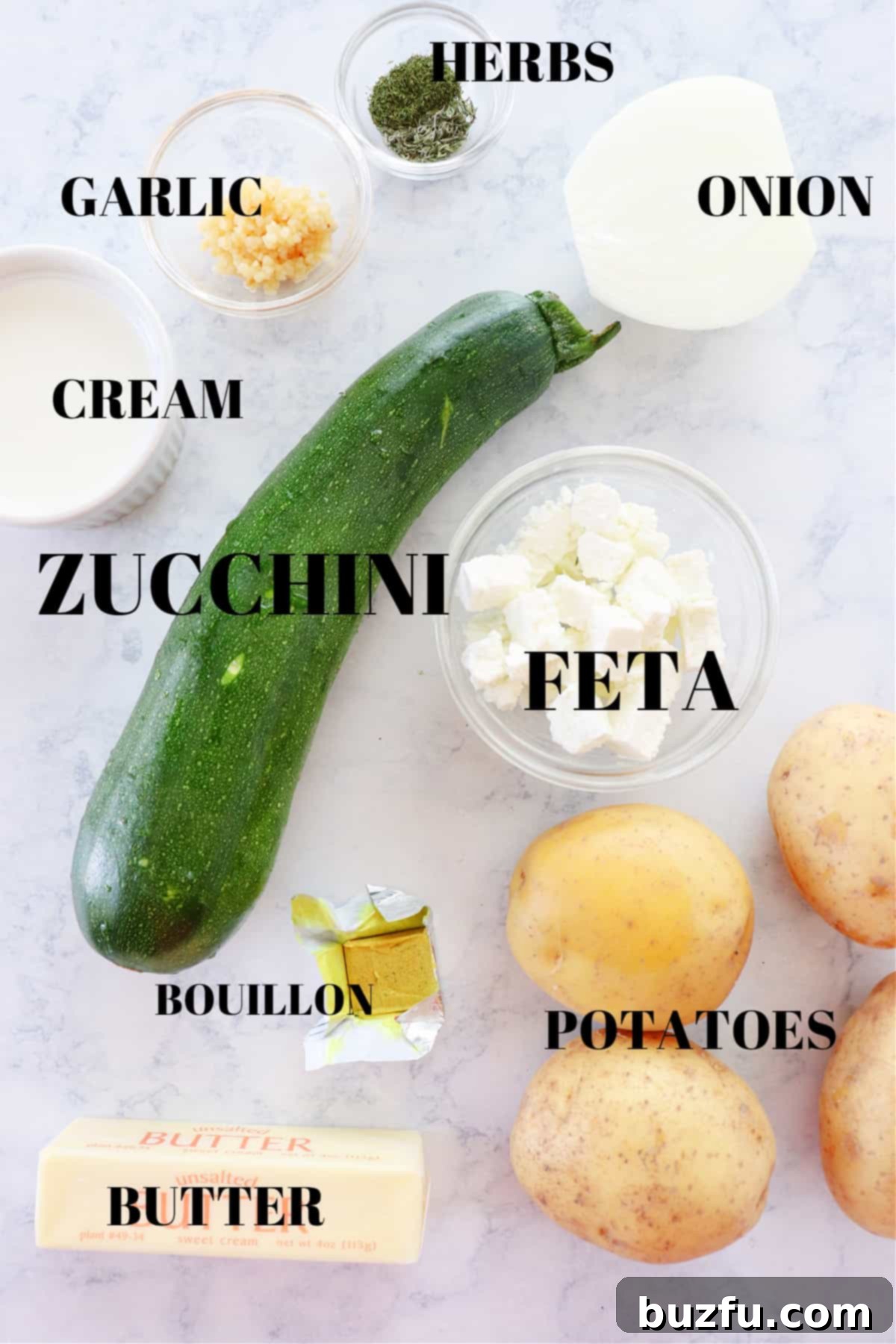 Assortment of fresh ingredients for zucchini soup, including zucchini, potatoes, onion, garlic, herbs, and feta, arranged on a marble cutting board.