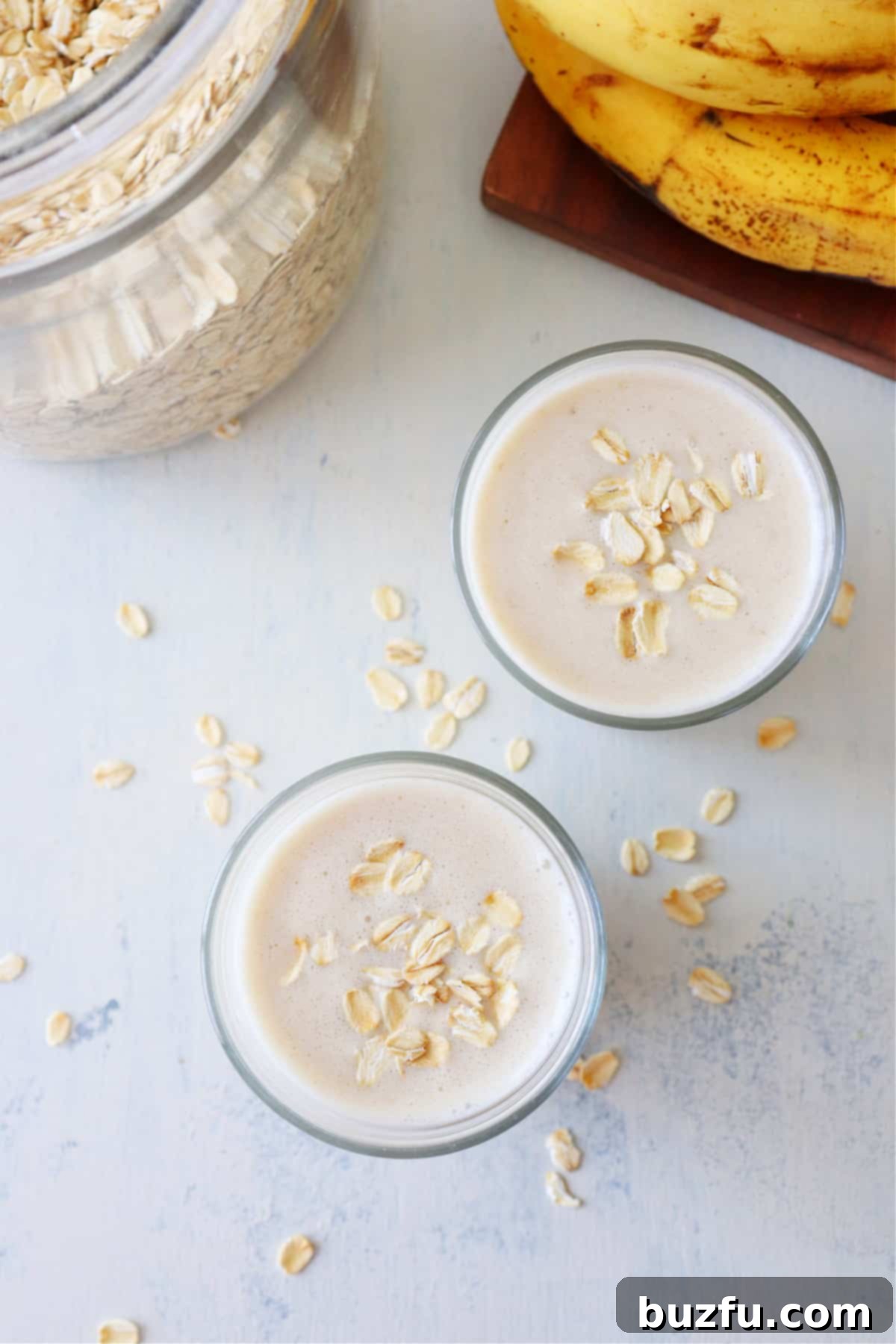 Two glasses of creamy oatmeal smoothie, artfully garnished with a sprinkle of rolled oats, presented on a minimalist white surface, highlighting a quick and healthy breakfast option.