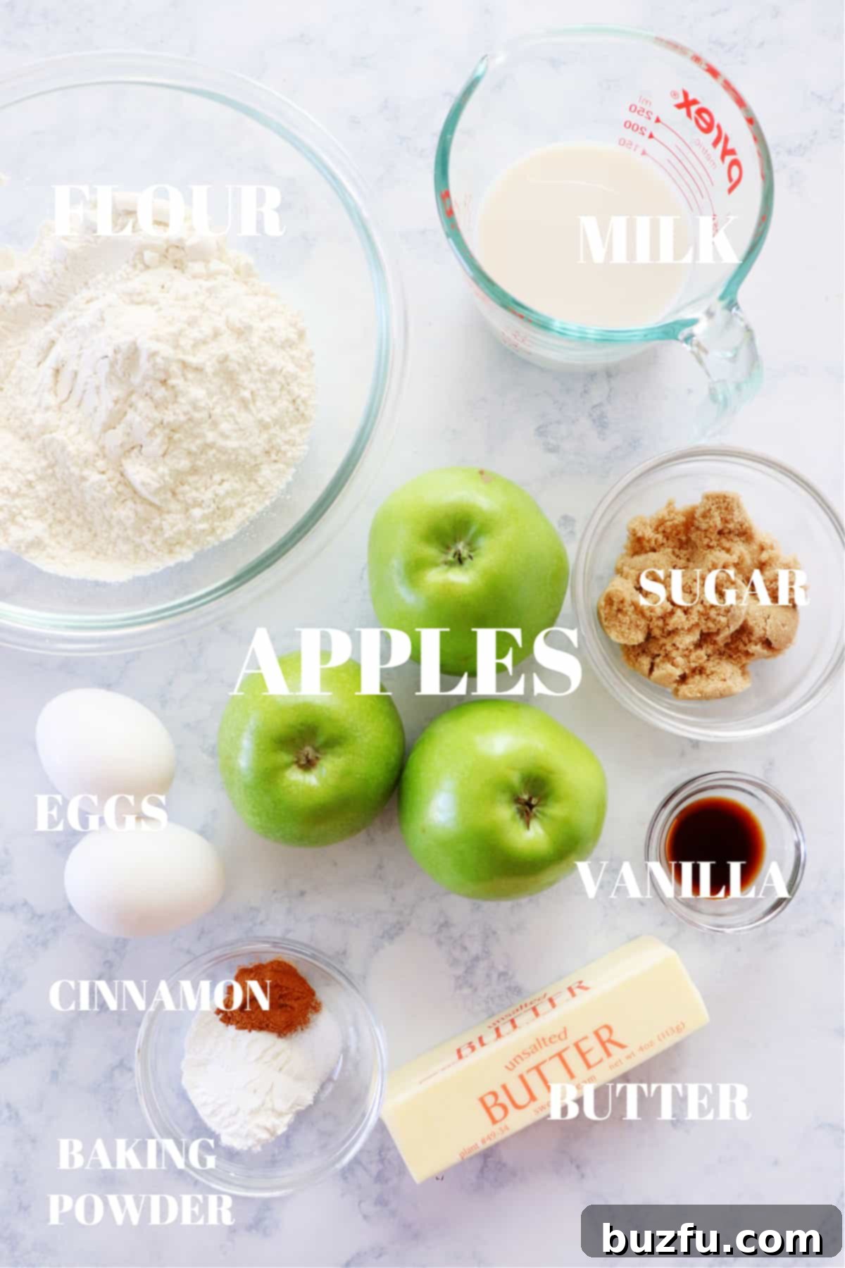 A selection of fresh apples, flour, sugar, eggs, and milk laid out on a marble board, ready for apple fritter preparation.