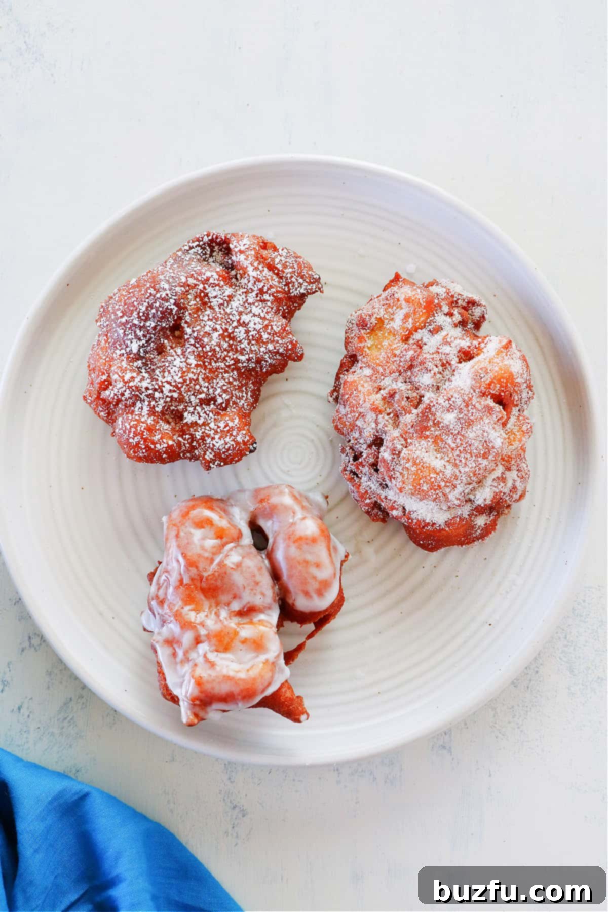 A plate showcasing three variations of apple fritters: one glazed, one dusted with powdered sugar, and one coated in cinnamon sugar.