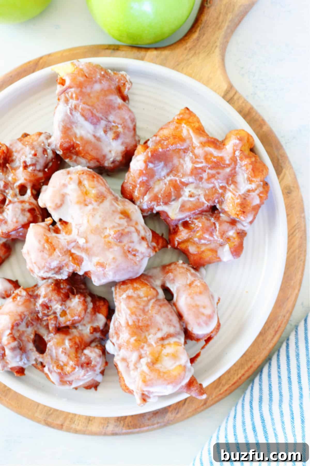 Several glazed apple fritters presented beautifully on a white plate with a rustic wooden board in the background, highlighting their delicious appearance.