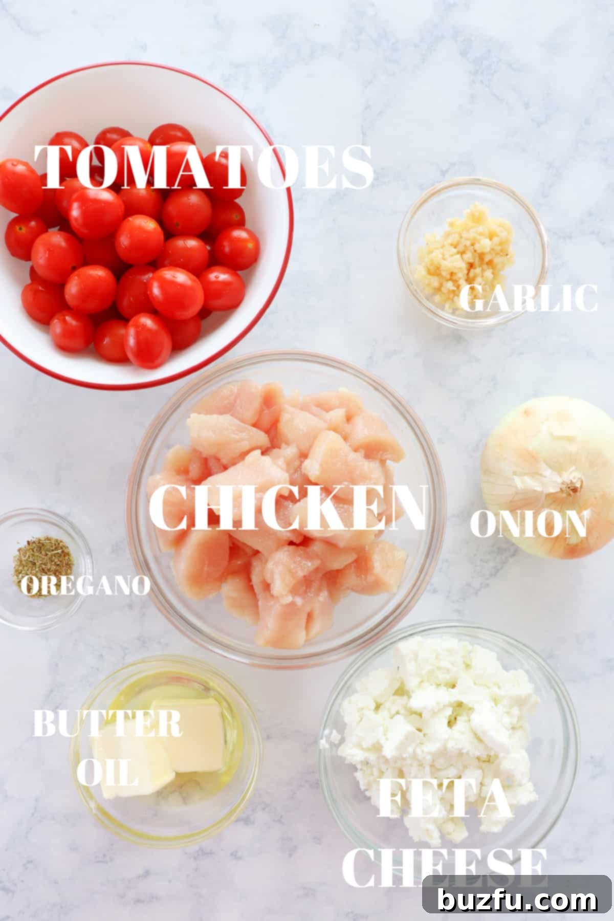 Fresh ingredients laid out on a marble board, including chicken breasts, cherry tomatoes, a block of feta cheese, an onion, garlic cloves, and seasonings, ready for the skillet.
