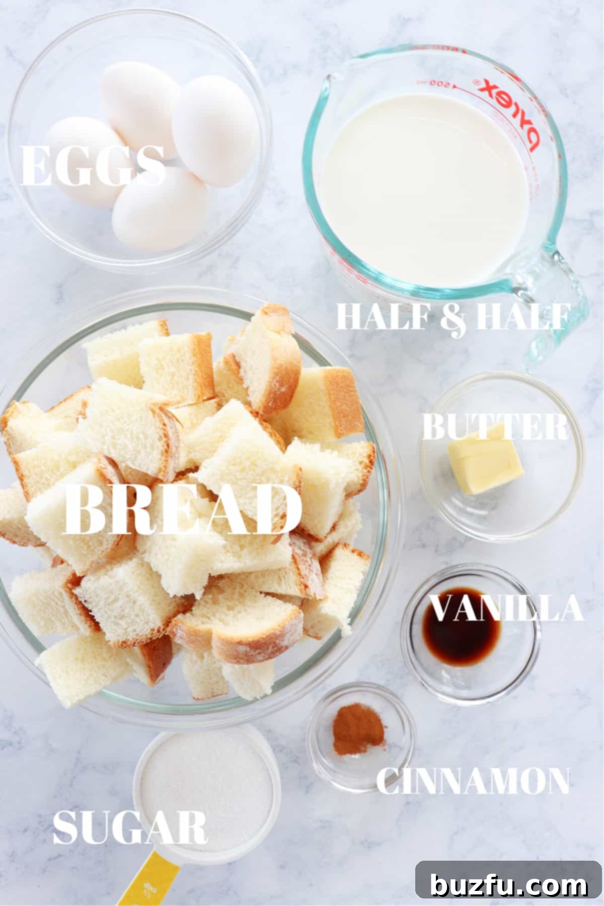 Ingredients for bread pudding displayed on a marble board, including cubed bread, eggs, milk, sugar, butter, vanilla, and cinnamon.