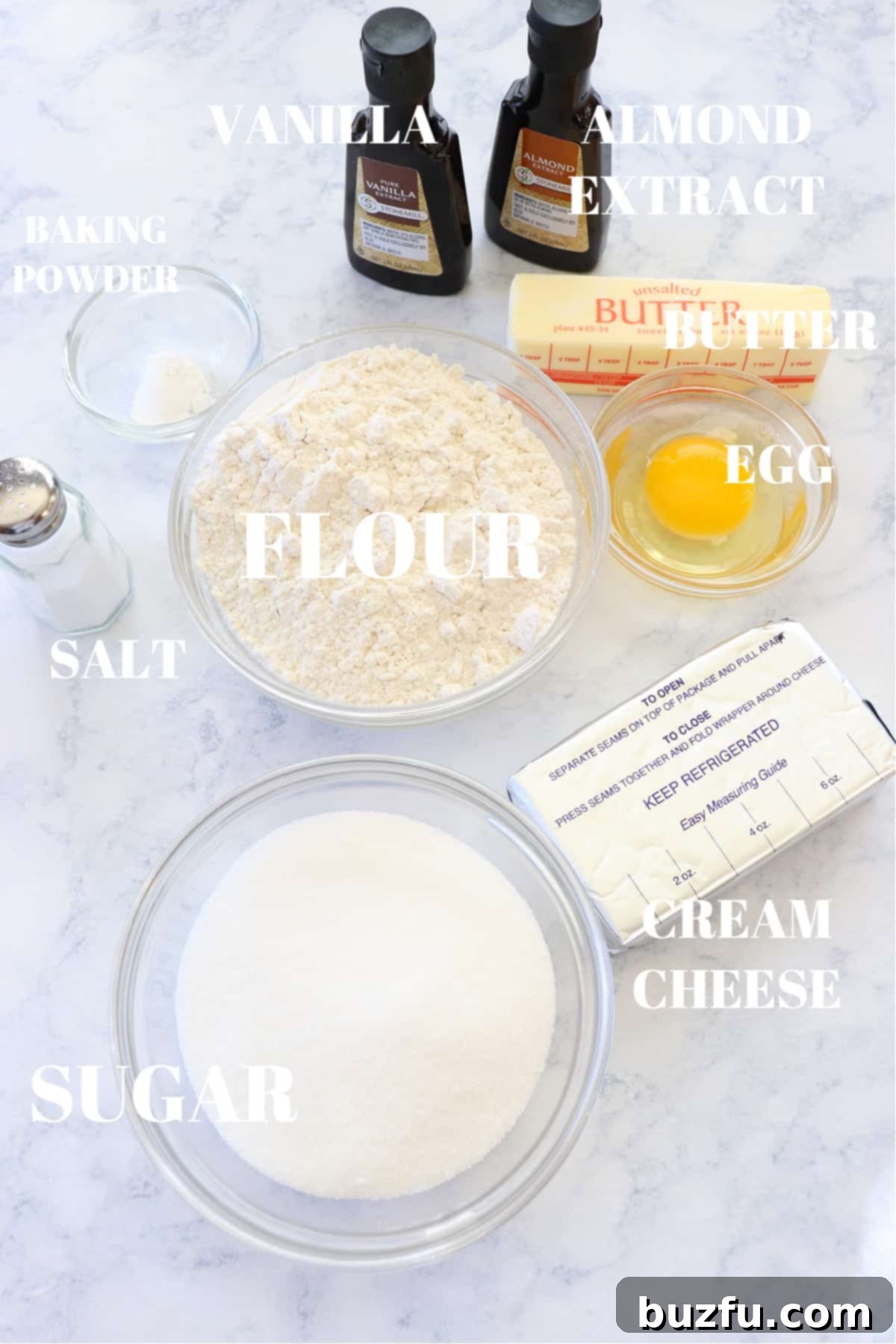 Various ingredients for cream cheese cookies laid out on a marble board, including butter, cream cheese, sugar, flour, and extracts.