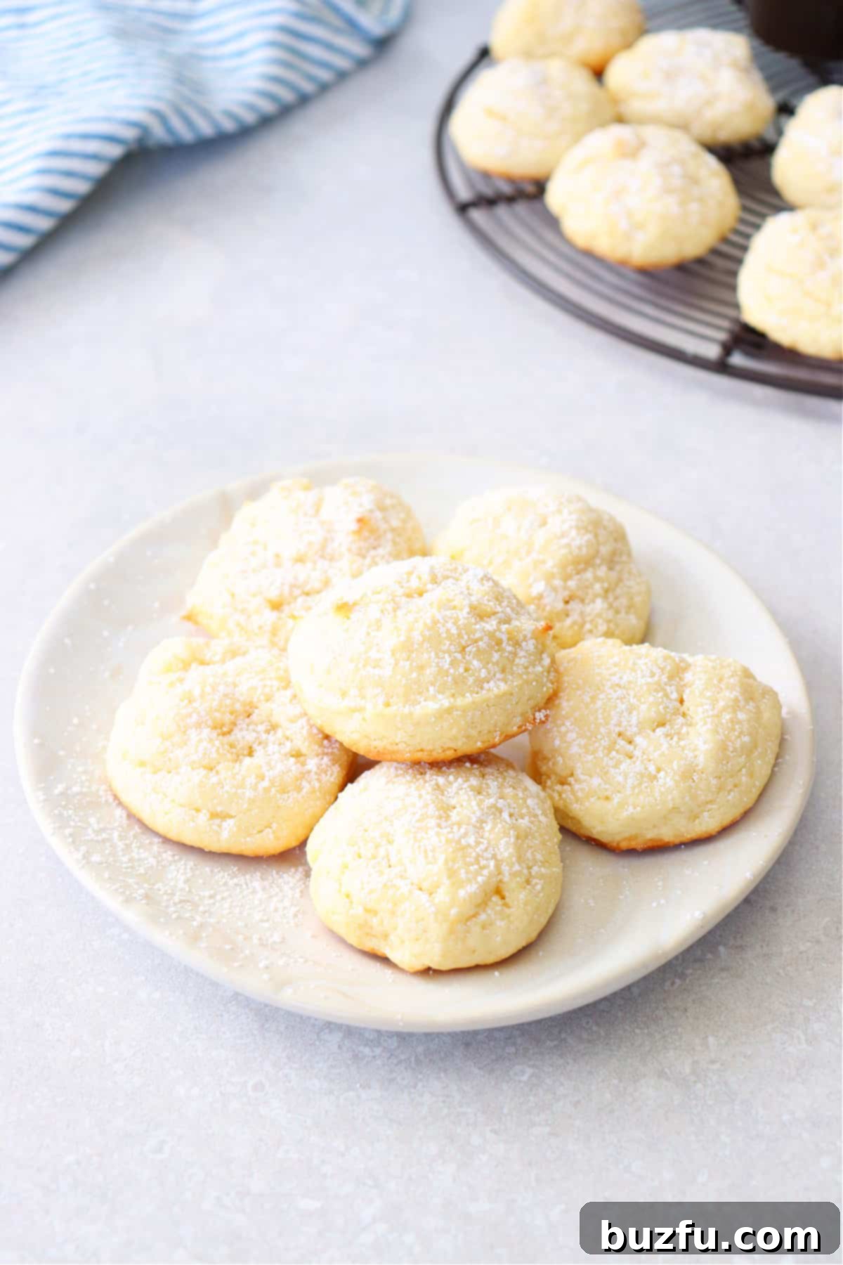 Beautifully arranged soft cream cheese cookies, lightly dusted with powdered sugar, on a cream-colored dessert plate, highlighting their delicate texture.