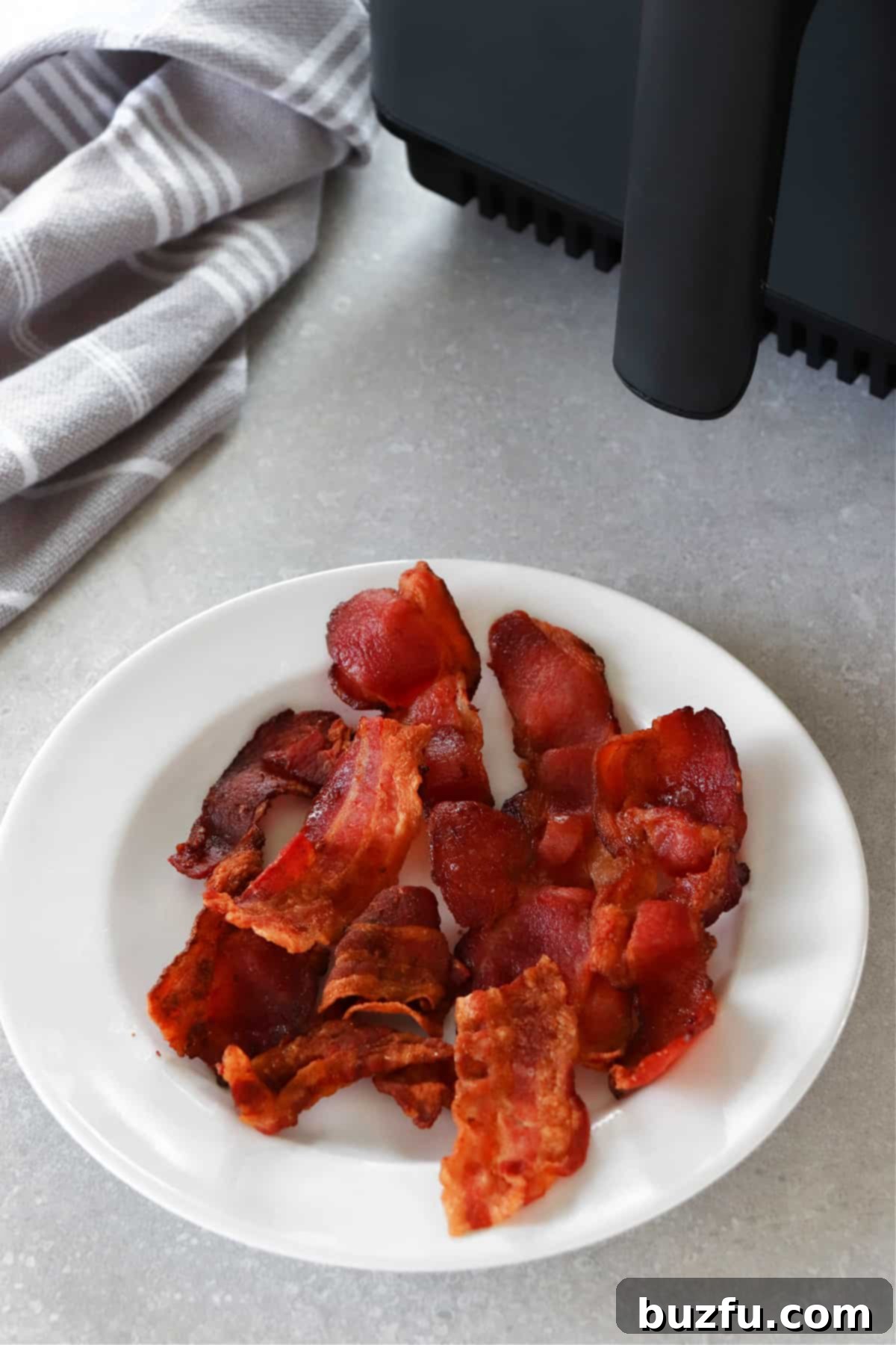 Crispy Air Fryer Bacon slices beautifully arranged on a white plate, positioned next to a modern air fryer in a kitchen setting. Golden brown and perfectly cooked.