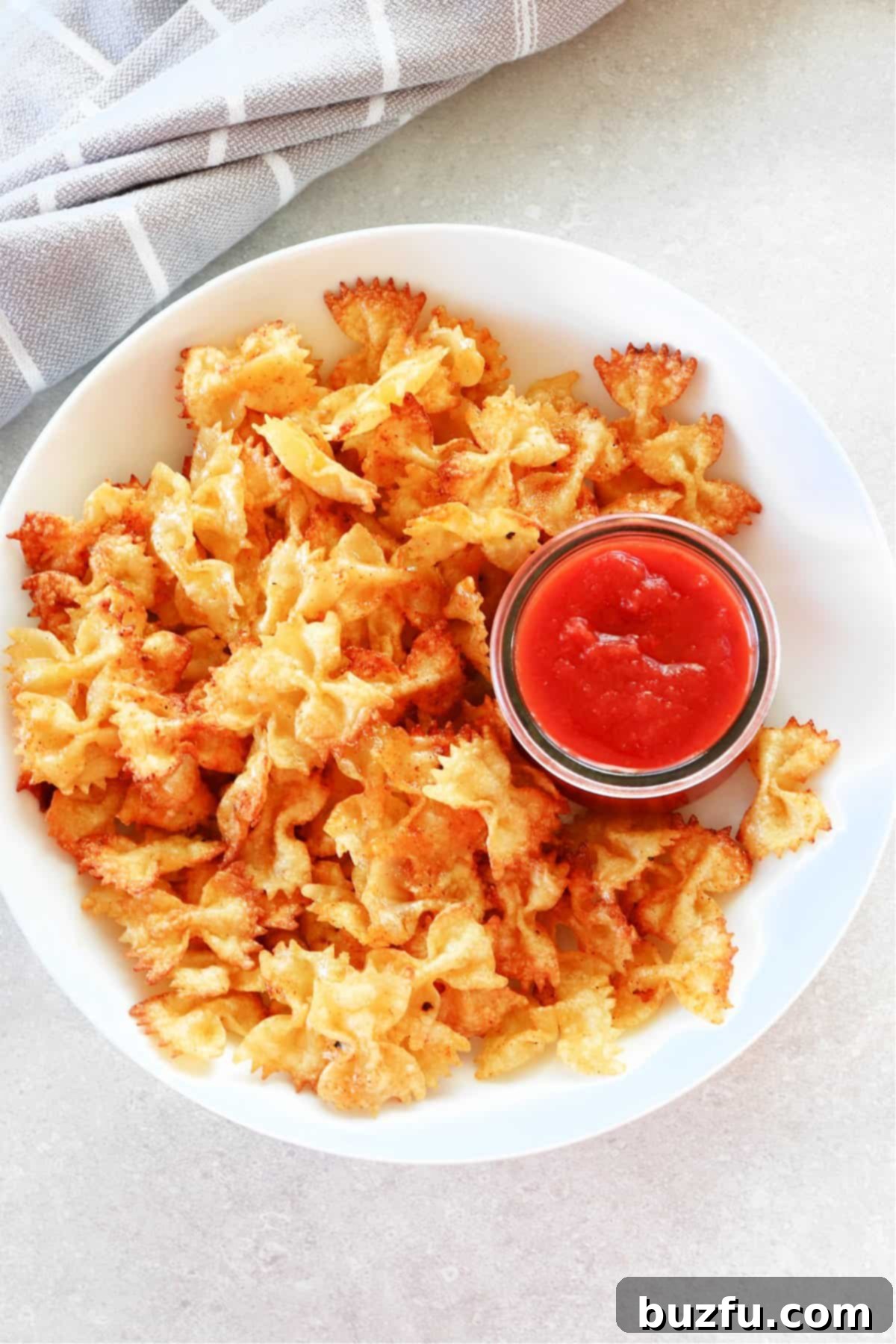 Delicious Air Fryer Pasta Chips. A white bowl filled with golden-brown air fryer pasta chips, accompanied by a small cup of marinara dipping sauce.