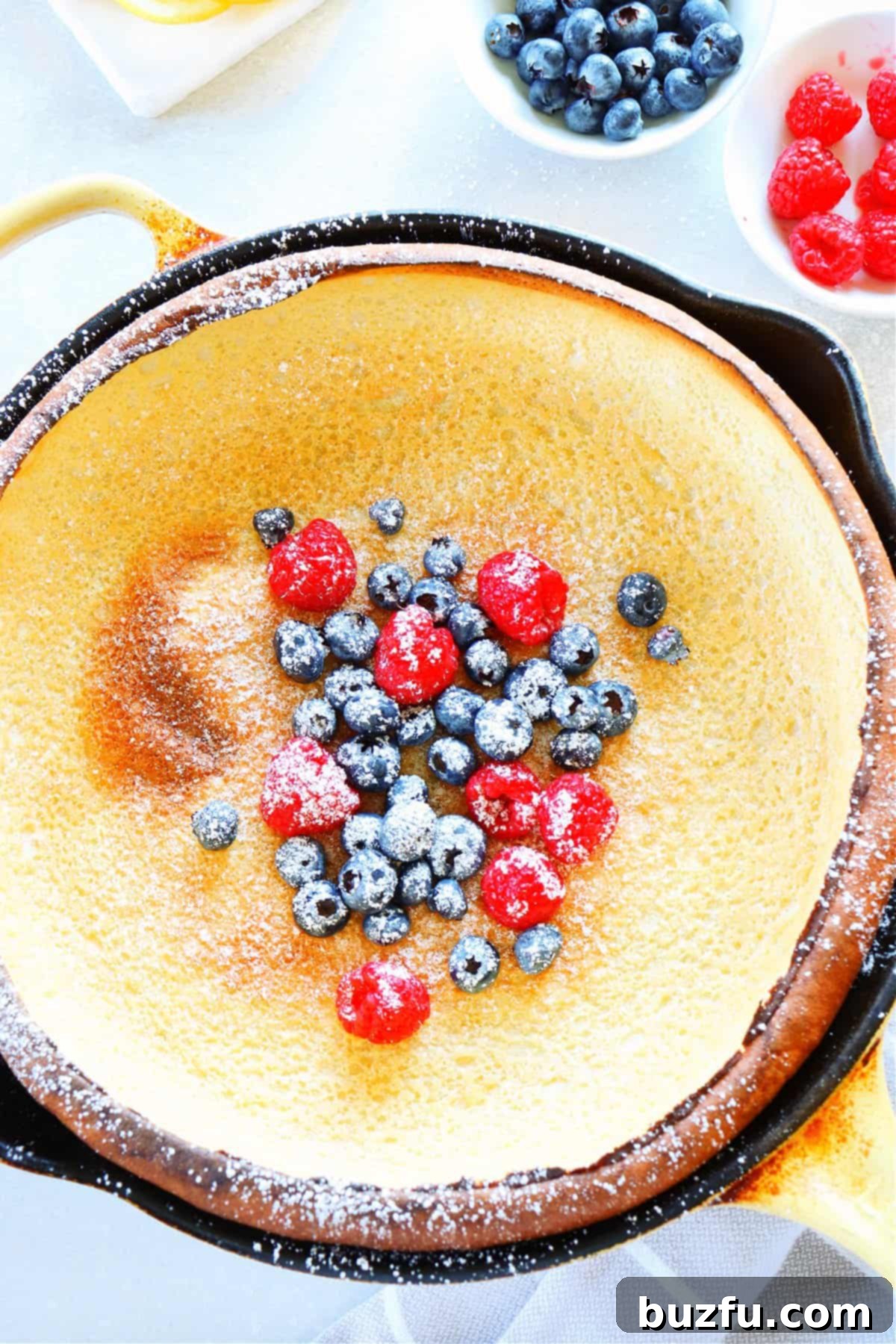 Delicious Dutch Baby pancake garnished with fresh blueberries and raspberries, dusted with powdered sugar, in a cast iron pan.