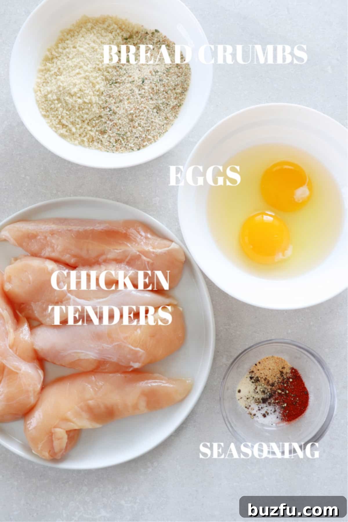 A selection of fresh ingredients for air fryer chicken tenders laid out on a wooden board, including chicken, eggs, breadcrumbs, and various seasonings.