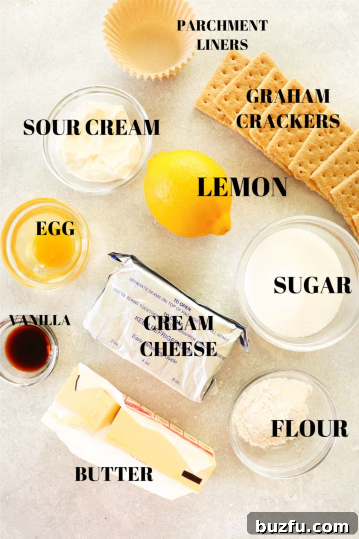 A collection of fresh ingredients for mini lemon cheesecakes laid out neatly on a rustic gray board, including cream cheese, fresh lemons, granulated sugar, vanilla extract, sour cream, all-purpose flour, eggs, graham crackers, and butter.