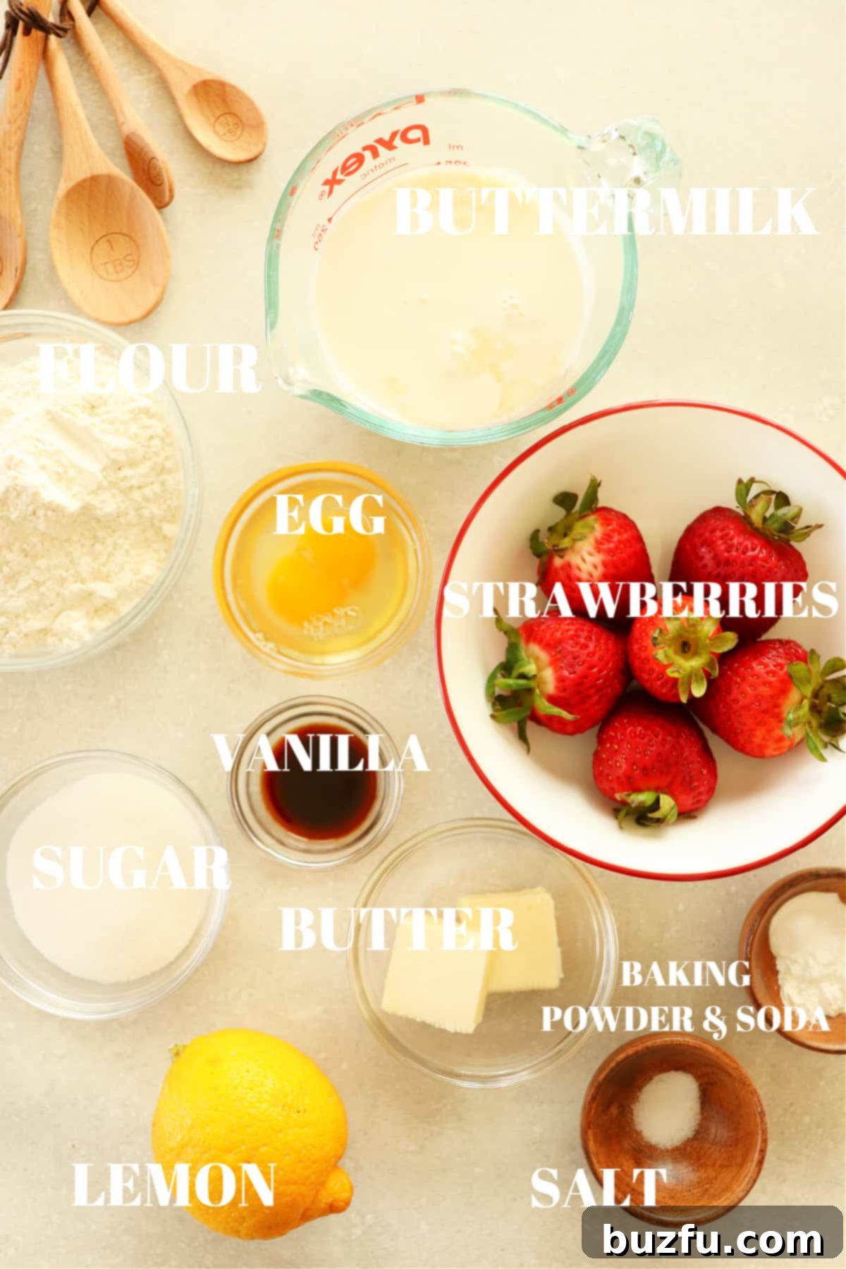 All ingredients for strawberry pancakes laid out on a gray board, including flour, baking soda, baking powder, sugar, salt, buttermilk, egg, butter, vanilla extract, lemon, and fresh strawberries.
