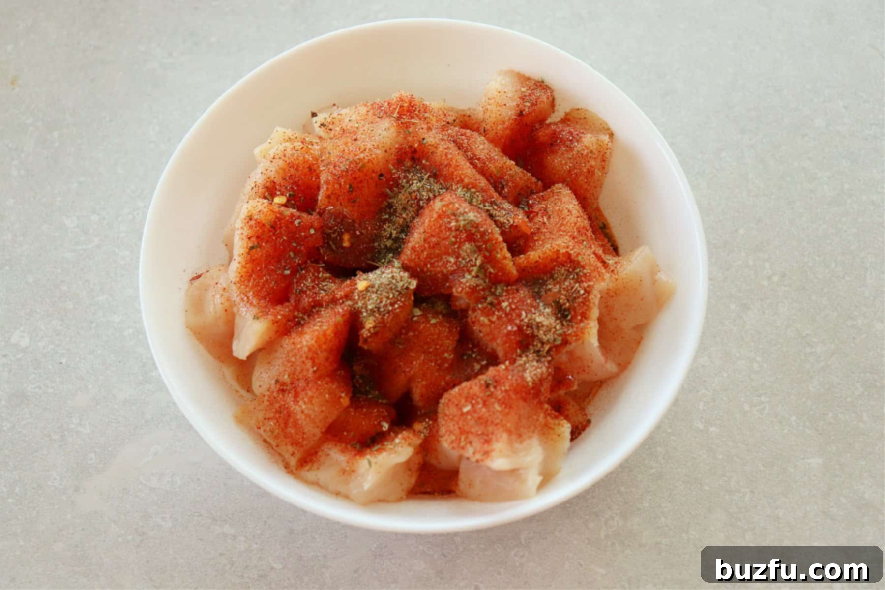 Raw chicken pieces uniformly cut and seasoned with spices and olive oil in a white mixing bowl.