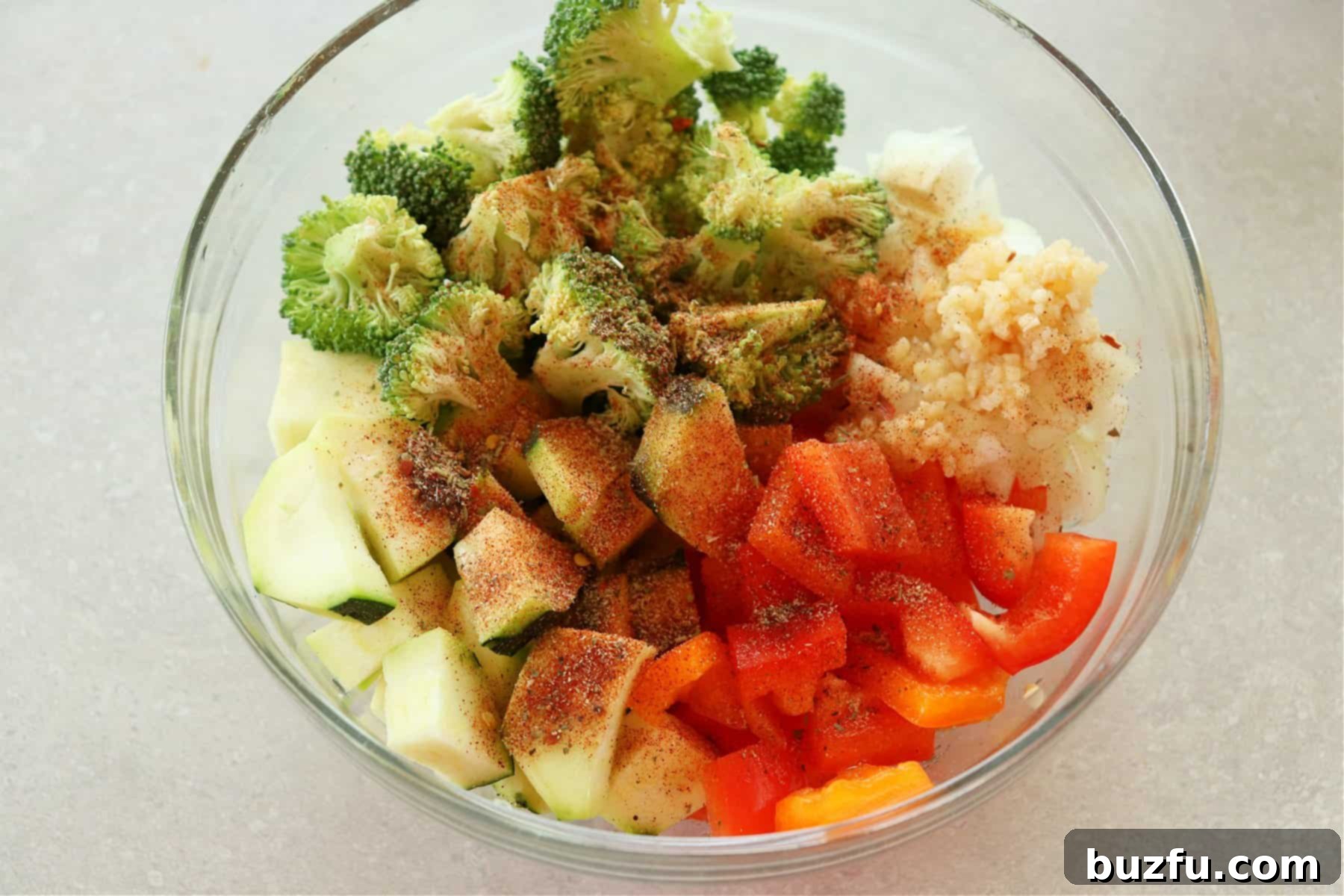 Chopped raw vegetables coated with olive oil and spices in a clear glass mixing bowl.