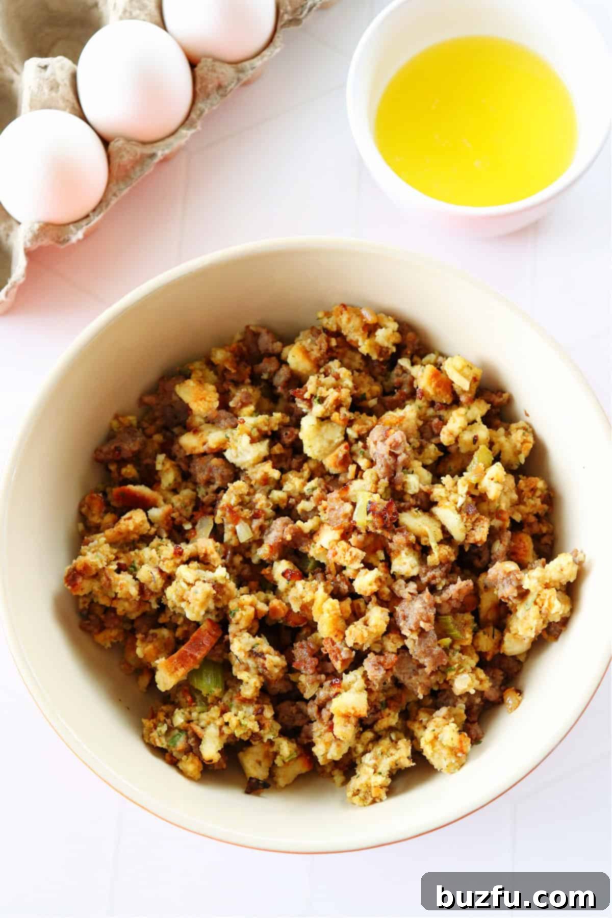 Savory Stuffing Cupcakes 3 Three core ingredients for stuffing muffins: a bowl of seasoned bread stuffing, a carton of fresh eggs, and a small dish of melted butter, all set on a clean white background.