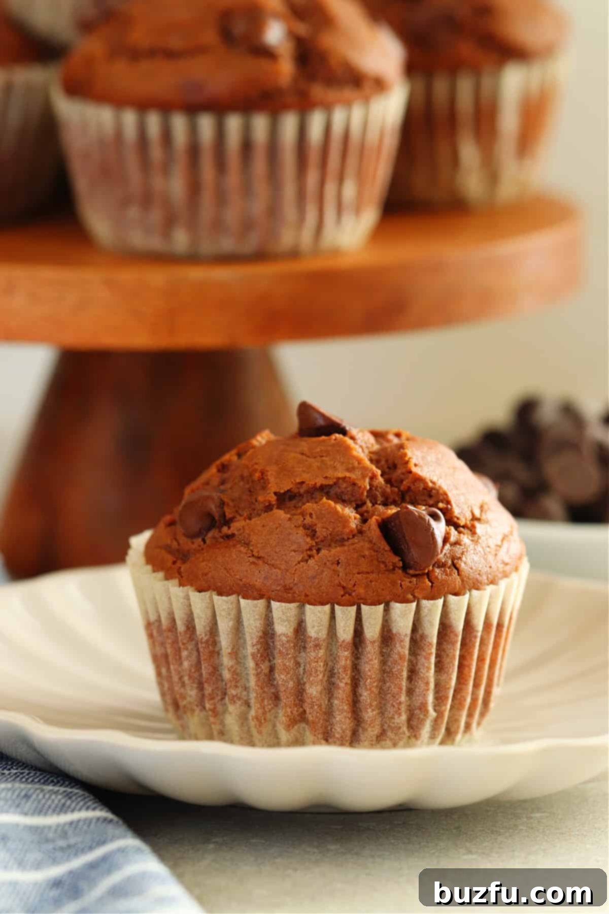Decadent Chocolate Muffins Made Simple 2 A perfectly baked, domed chocolate muffin sits on a small white plate, with a rustic wooden cake stand laden with more warm muffins blurred in the background, hinting at abundant deliciousness.