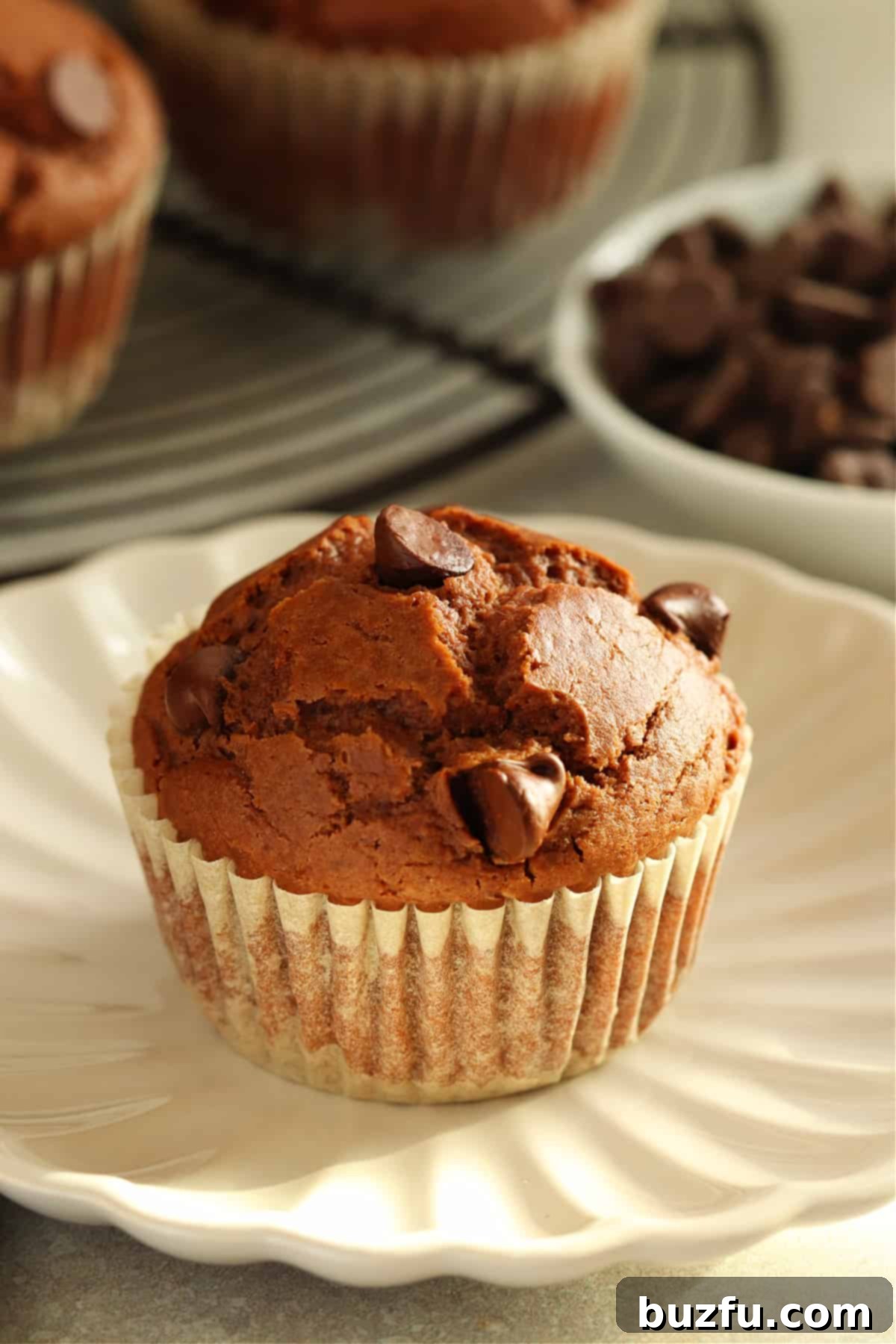 Decadent Chocolate Muffins Made Simple 7 A perfectly baked chocolate muffin, still warm in its parchment wrapper, rests on a small, elegant plate, with more enticing muffins softly blurred in the background.