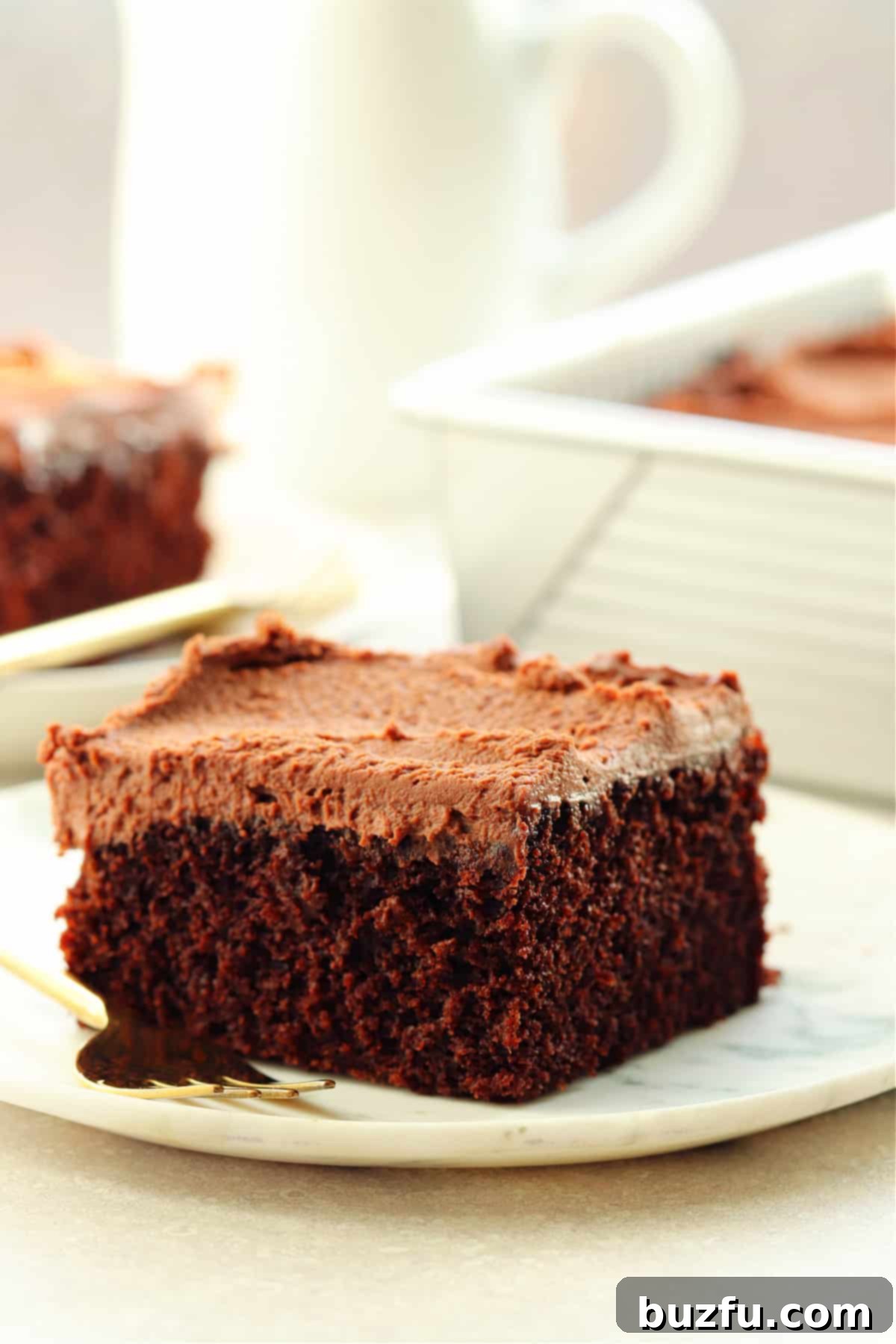 Decadent Eggless Chocolate Delight 7 One piece of chocolate cake with frosting on a white plate with a pan with the cake in the background.