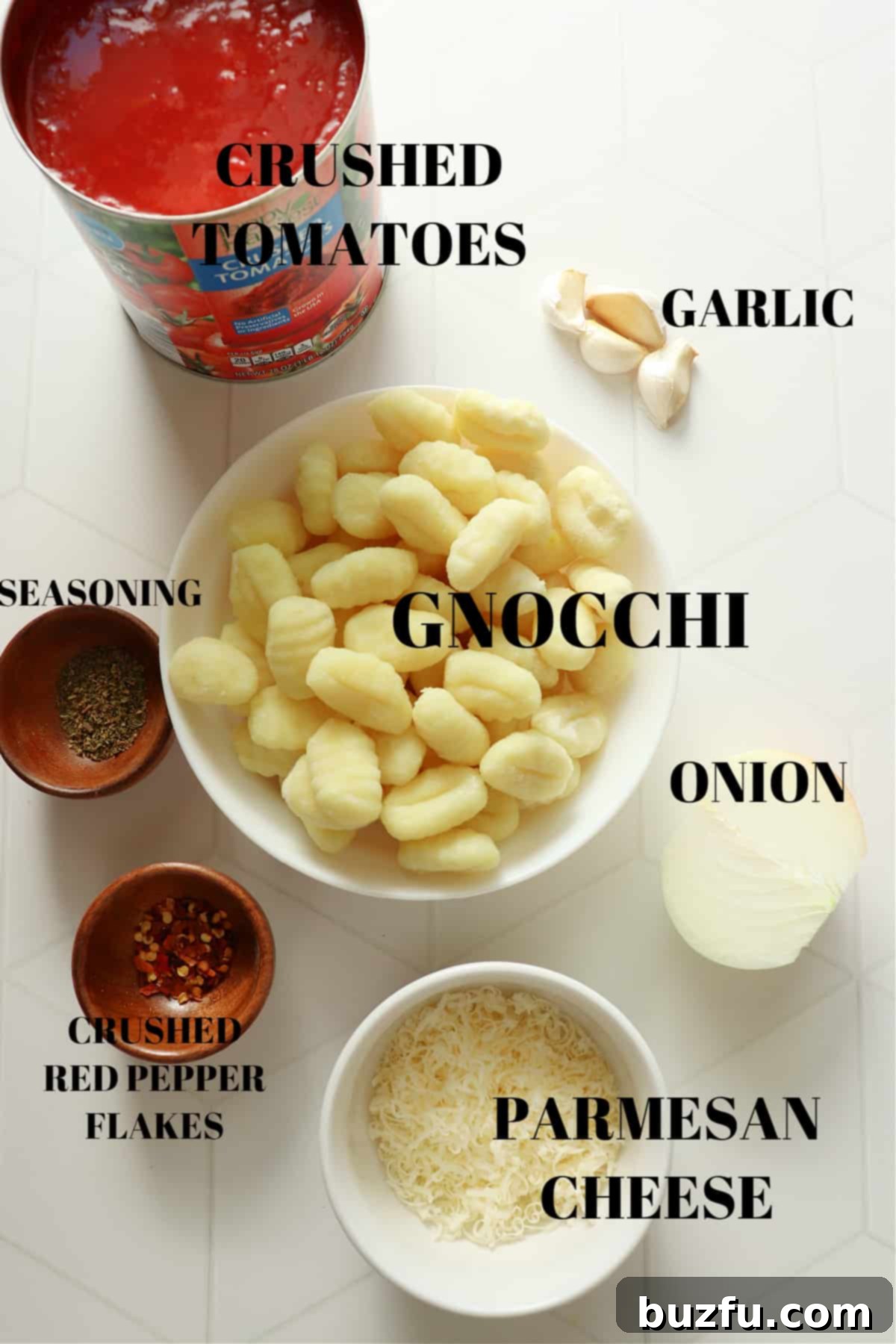 Gnocchi, crushed tomatoes, Parmesan, onion, garlic cloves and seasoning in bowls on a white tile board.