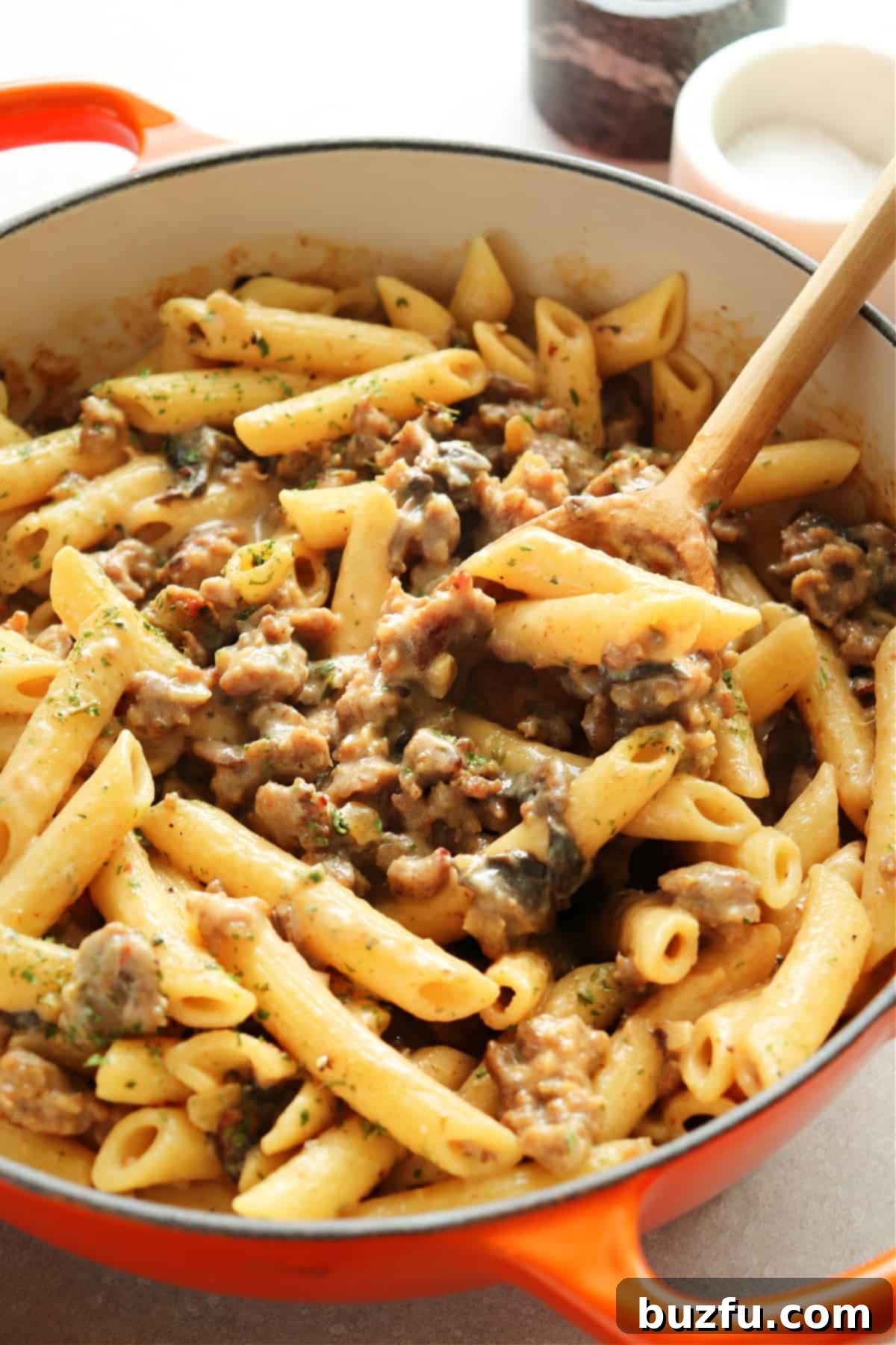 Penne and sausage in an orange Dutch oven on a gray board.
