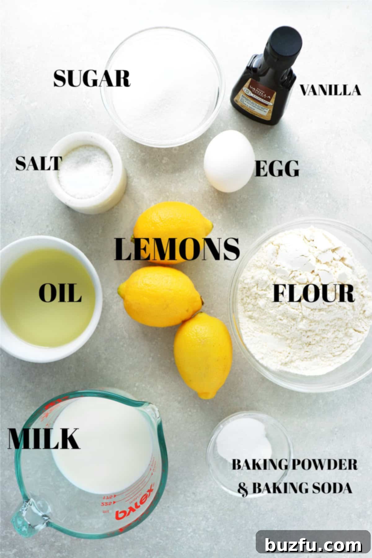 A flat lay of all the fresh and dry ingredients required for baking a lemon cake, neatly arranged on a rustic gray board.