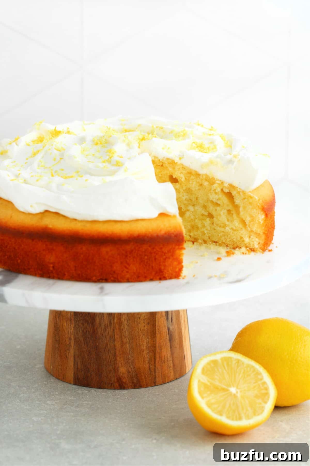 A beautiful round lemon cake with a generous layer of whipped cream, resting elegantly on a marble cake stand with fresh lemons scattered nearby.
