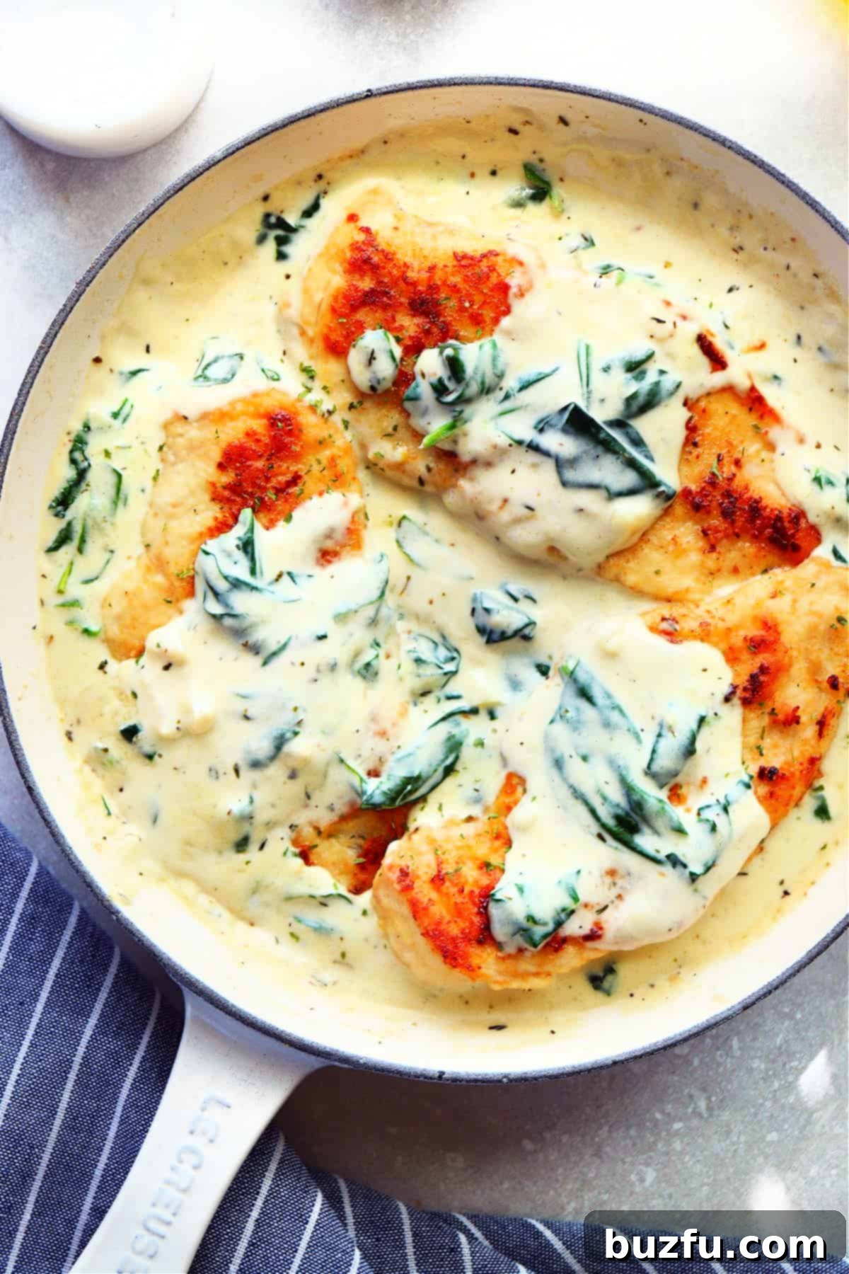 A stunning close-up of three cooked chicken breasts, beautifully coated in a creamy spinach sauce, presented in an elegant enameled Le Creuset skillet.