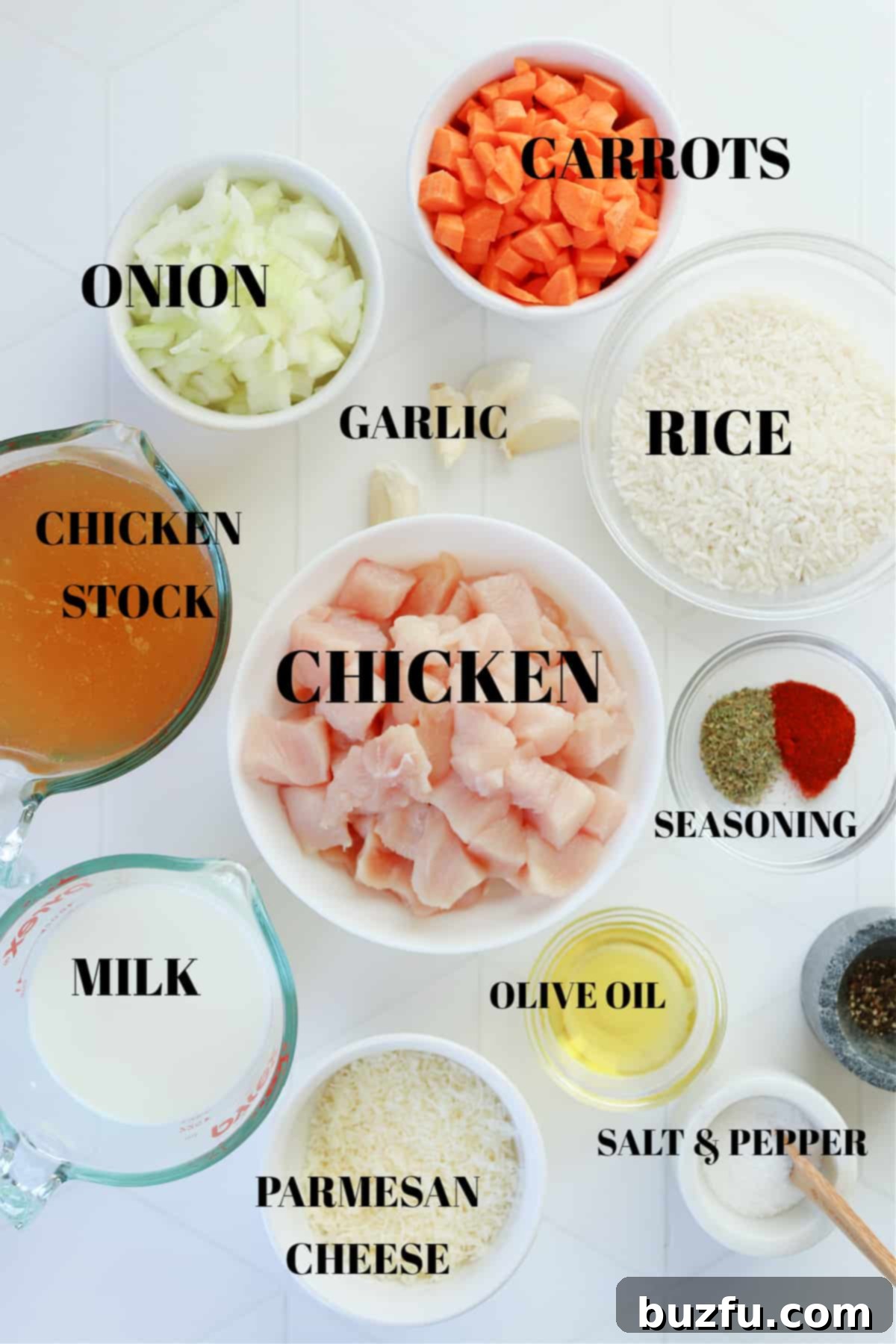 A collection of fresh ingredients for creamy chicken and rice, including chicken breasts, olive oil, spices, garlic, onion, carrots, rice, broth, milk, and Parmesan cheese, arranged in bowls on a white tile surface.
