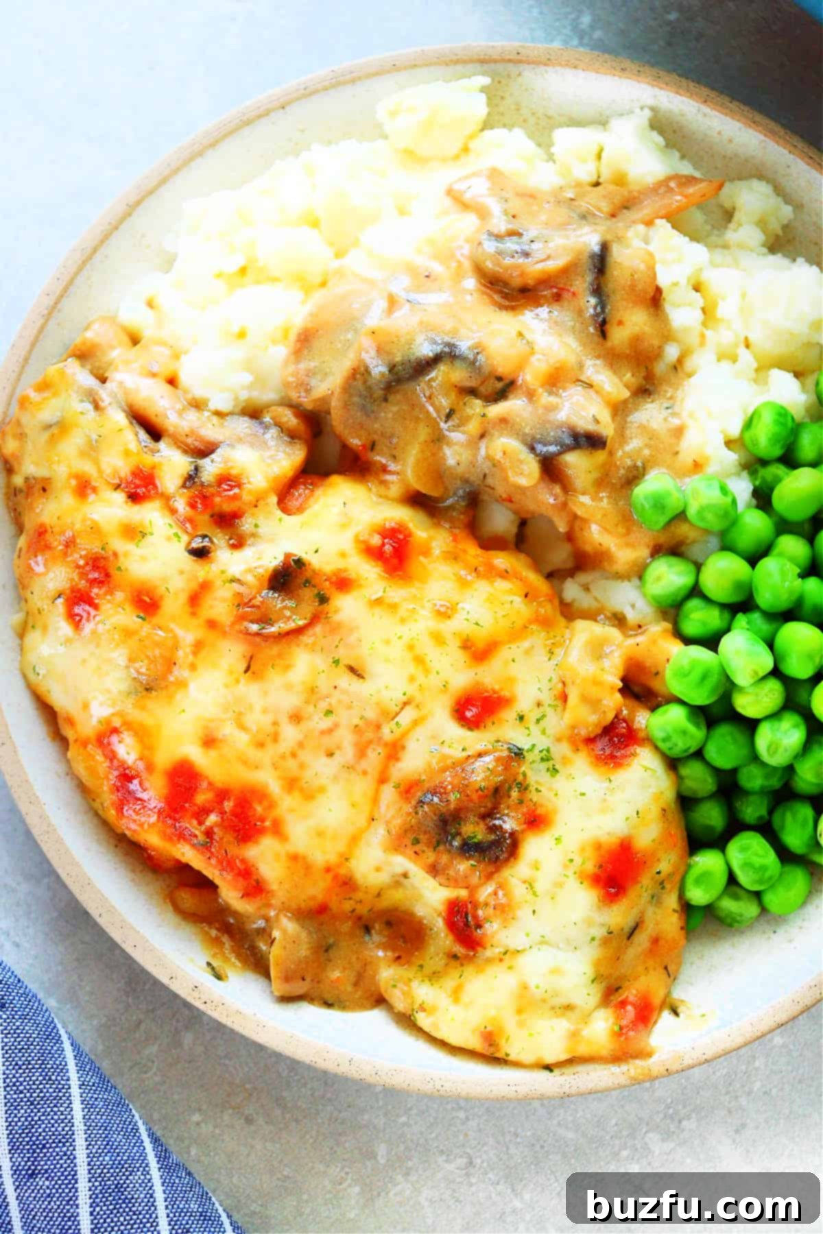 Chicken breast with melted cheese, mashed potatoes with mushroom gravy and steamed green peas on a dinner plate.