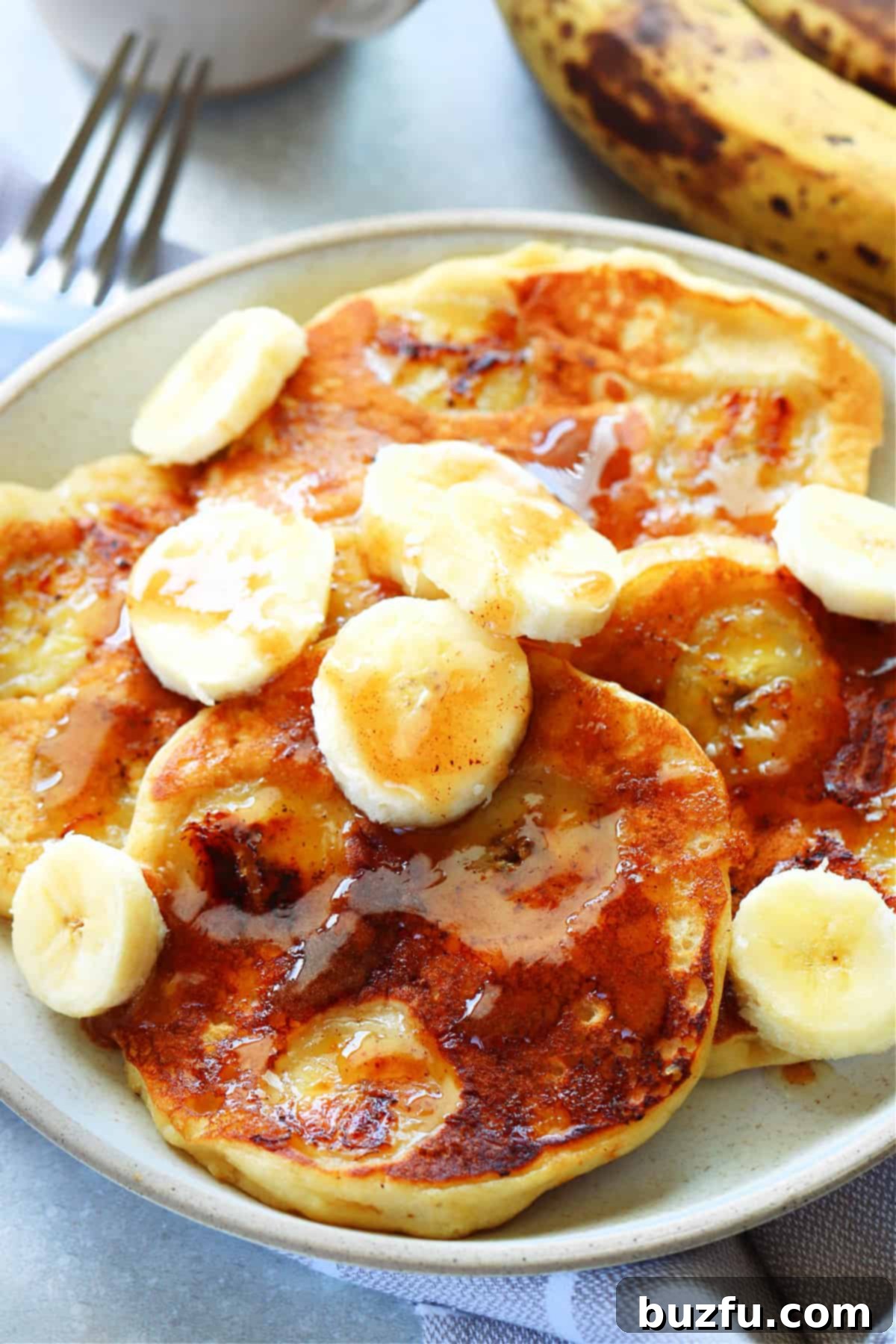 Golden Banana Pancakes 2 Four golden brown banana pancakes, generously drizzled with rich brown sugar butter and garnished with fresh banana slices on a pristine white plate, accompanied by a fork, ready to be enjoyed.