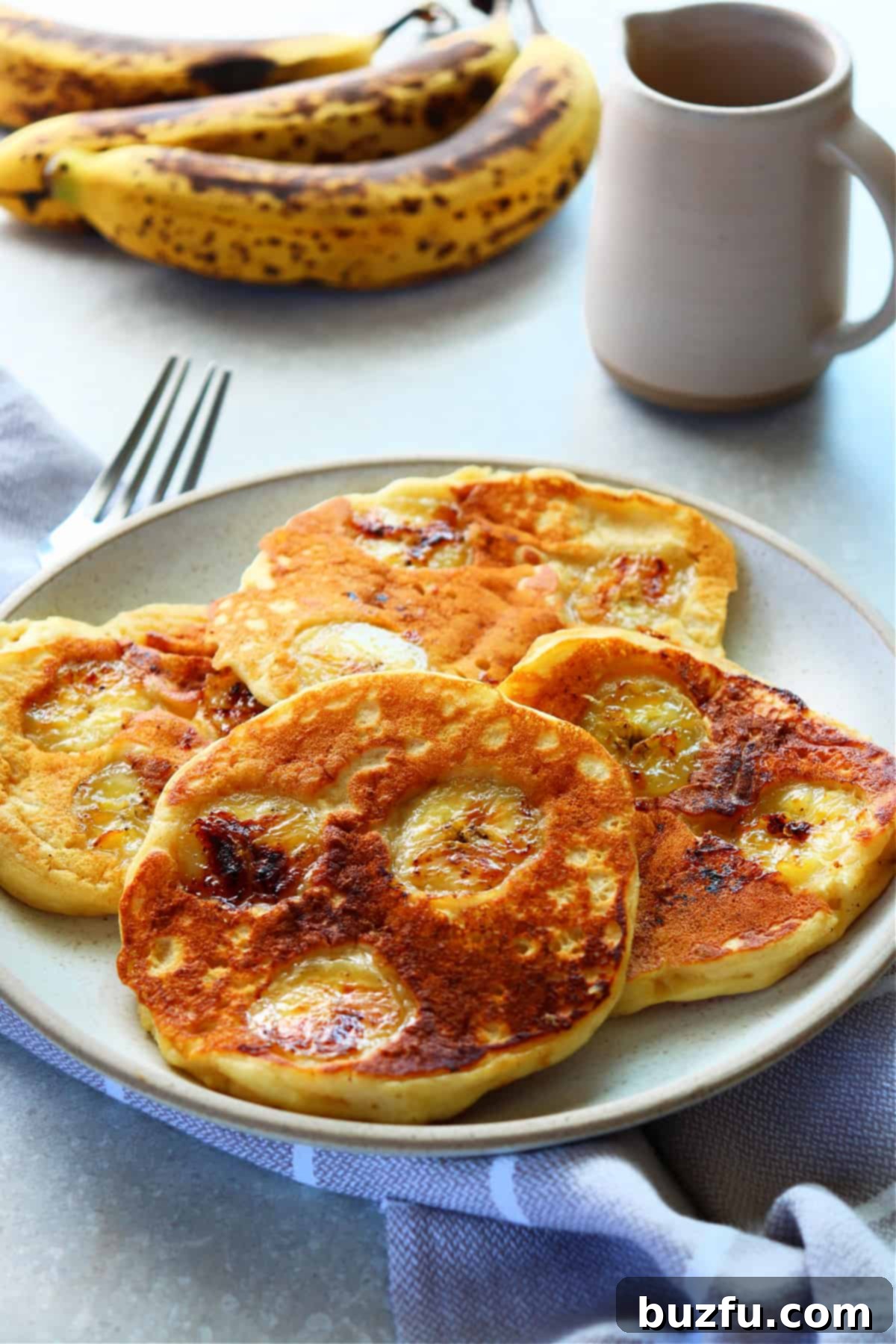 Golden Banana Pancakes 6 Four banana pancakes on a clean white plate with a fork resting on a folded napkin, suggesting a delicious and inviting breakfast.