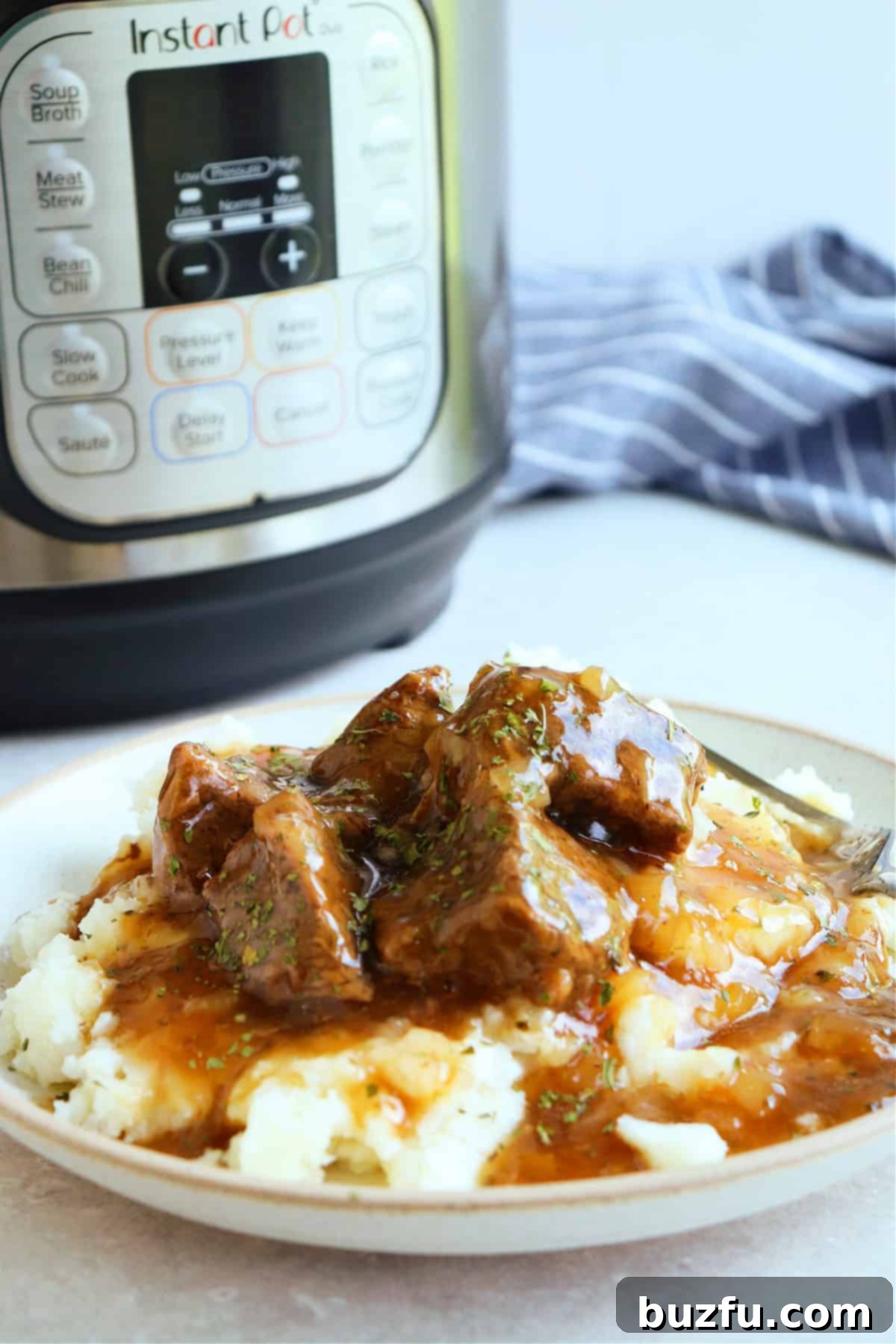 Week Six Meal Plan 7 A side view of tender beef tips generously covered in a rich gravy, served over a bed of creamy mashed potatoes on a white plate, with the Instant Pot subtly blurred in the background.