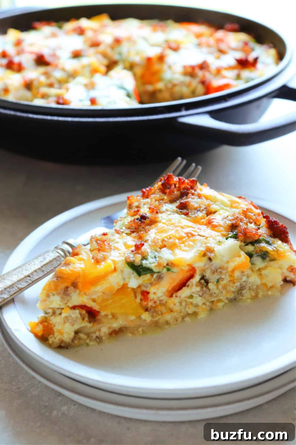 A photo of a slice of quiche on a plate with a fork, with the whole quiche in a cast iron pan in the background.