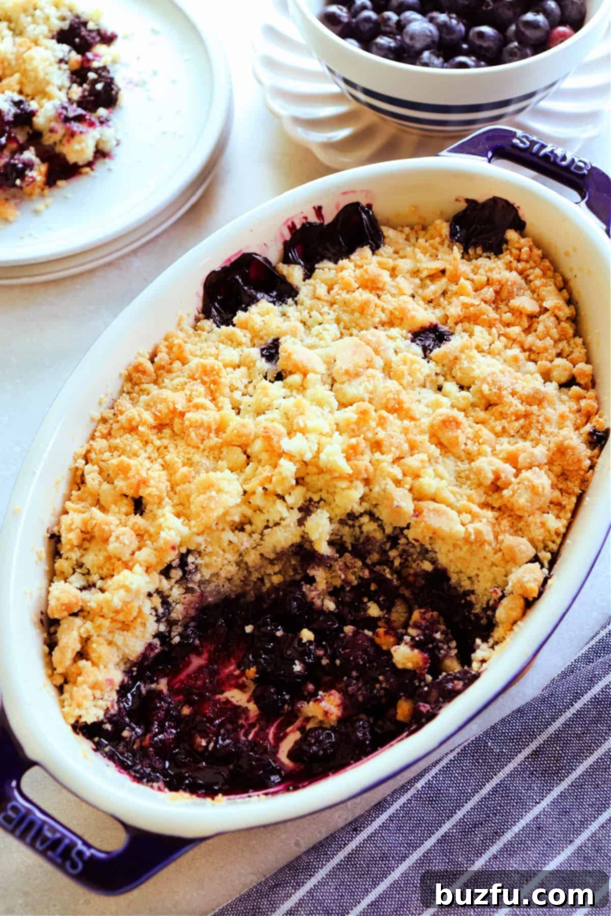 Blueberry crumble in a blue stoneware baking dish with a serving missing, with blue striped towel next to it, and blueberries in a bowl behind it.